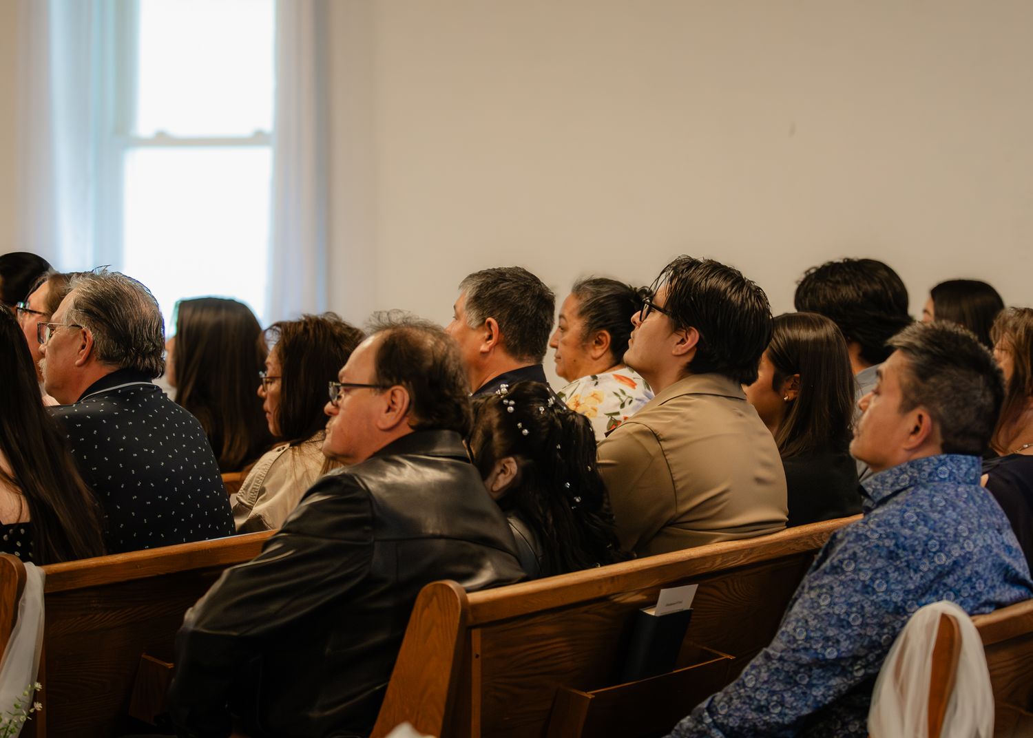 Wedding guests seated in wooden pews watching ceremony in bright white chapel.