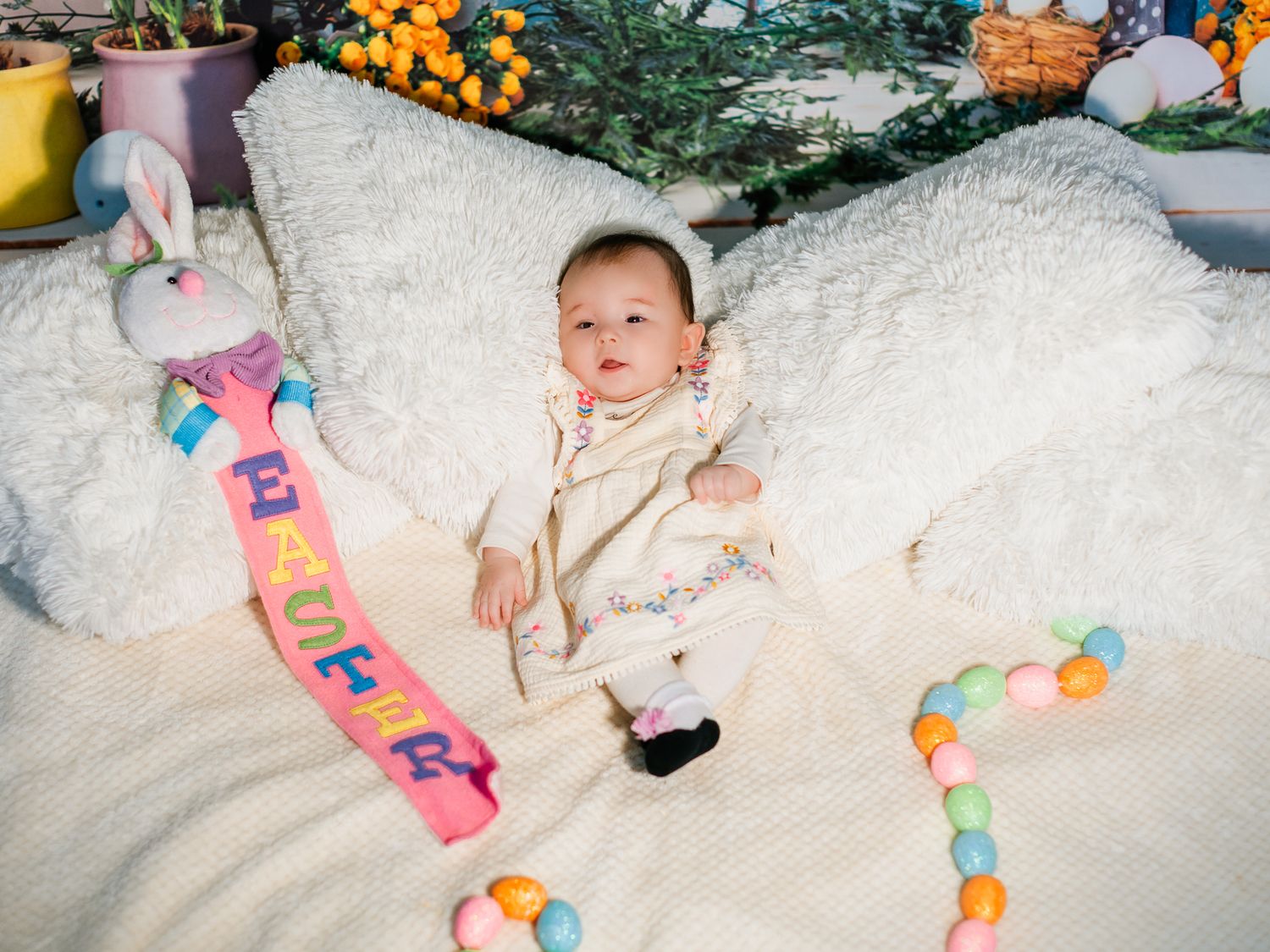 Baby in cream outfit posed on white fluffy blanket with Easter bunny, eggs, and colorful EASTER banner.