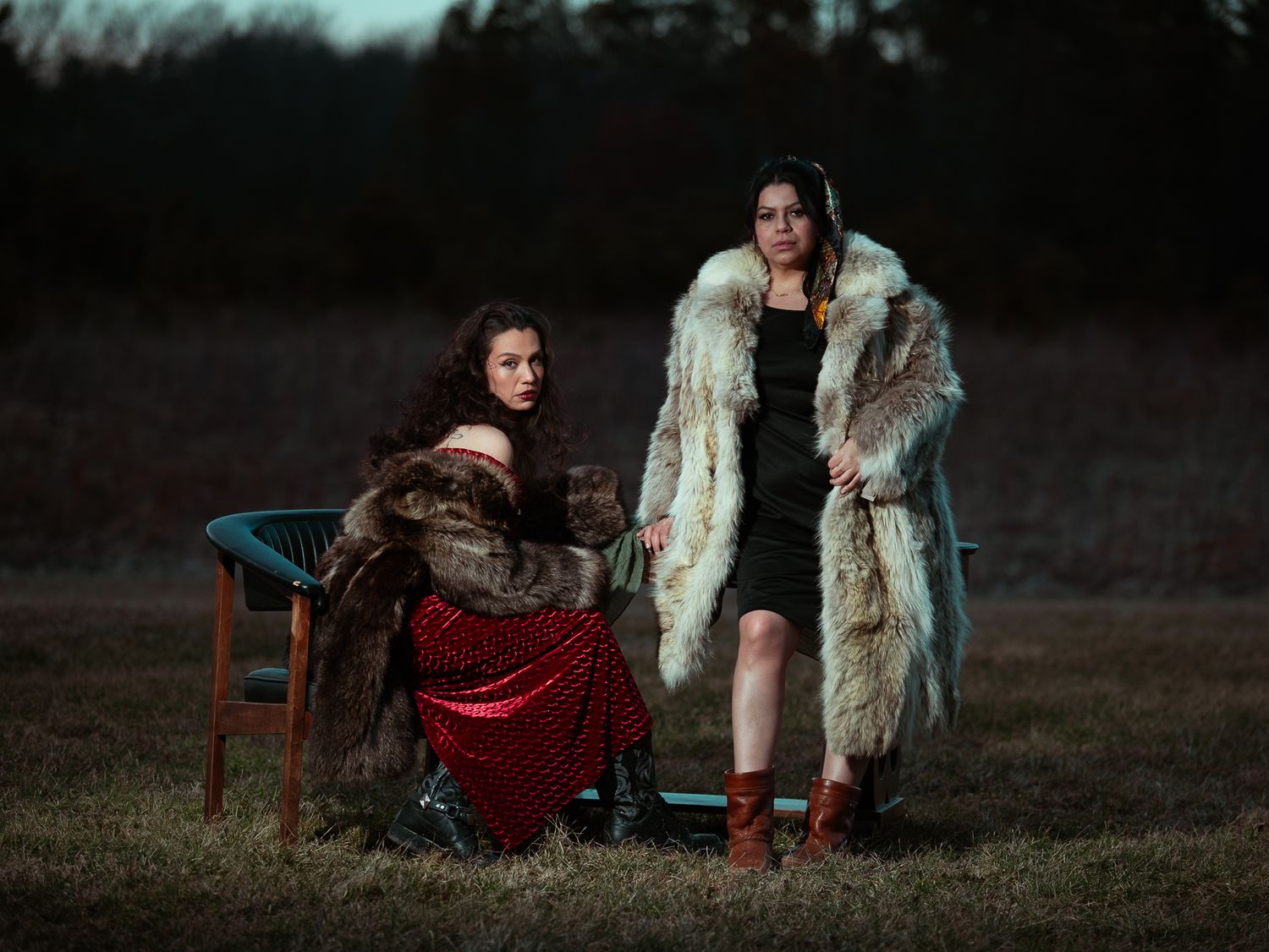 Two women in fur coats pose dramatically outdoors at dusk in a moody, atmospheric fashion portrait.
