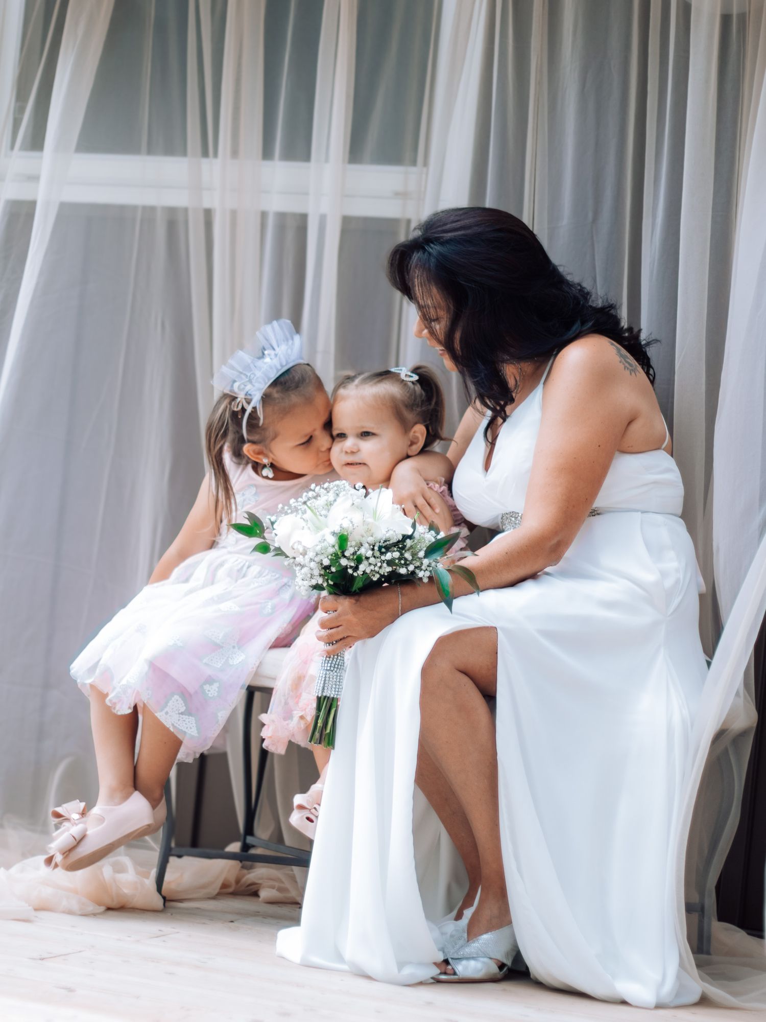 Bride in white gown sits with two young flower girls in pink dresses holding white bouquet.