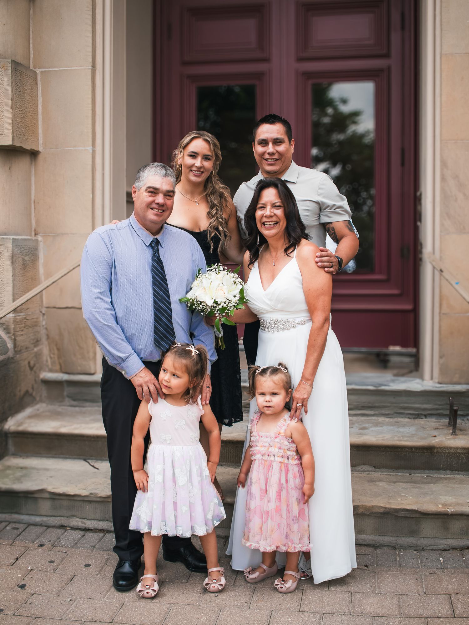 Family poses on steps outside a building during a wedding celebration with two young girls in pink dresses.