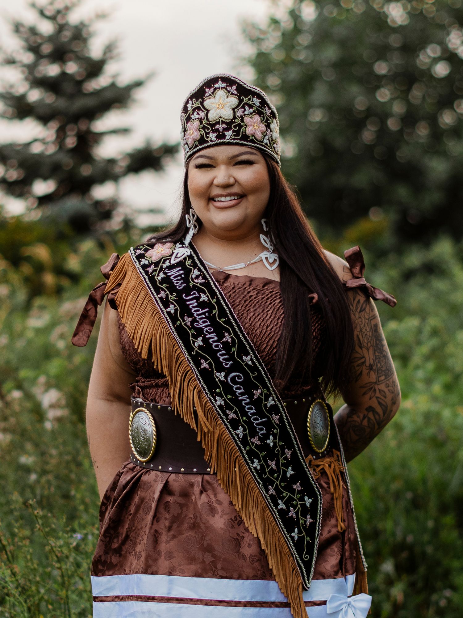 Smiling woman in traditional Indigenous regalia with crown and Miss Indigenous Canada sash outdoors.