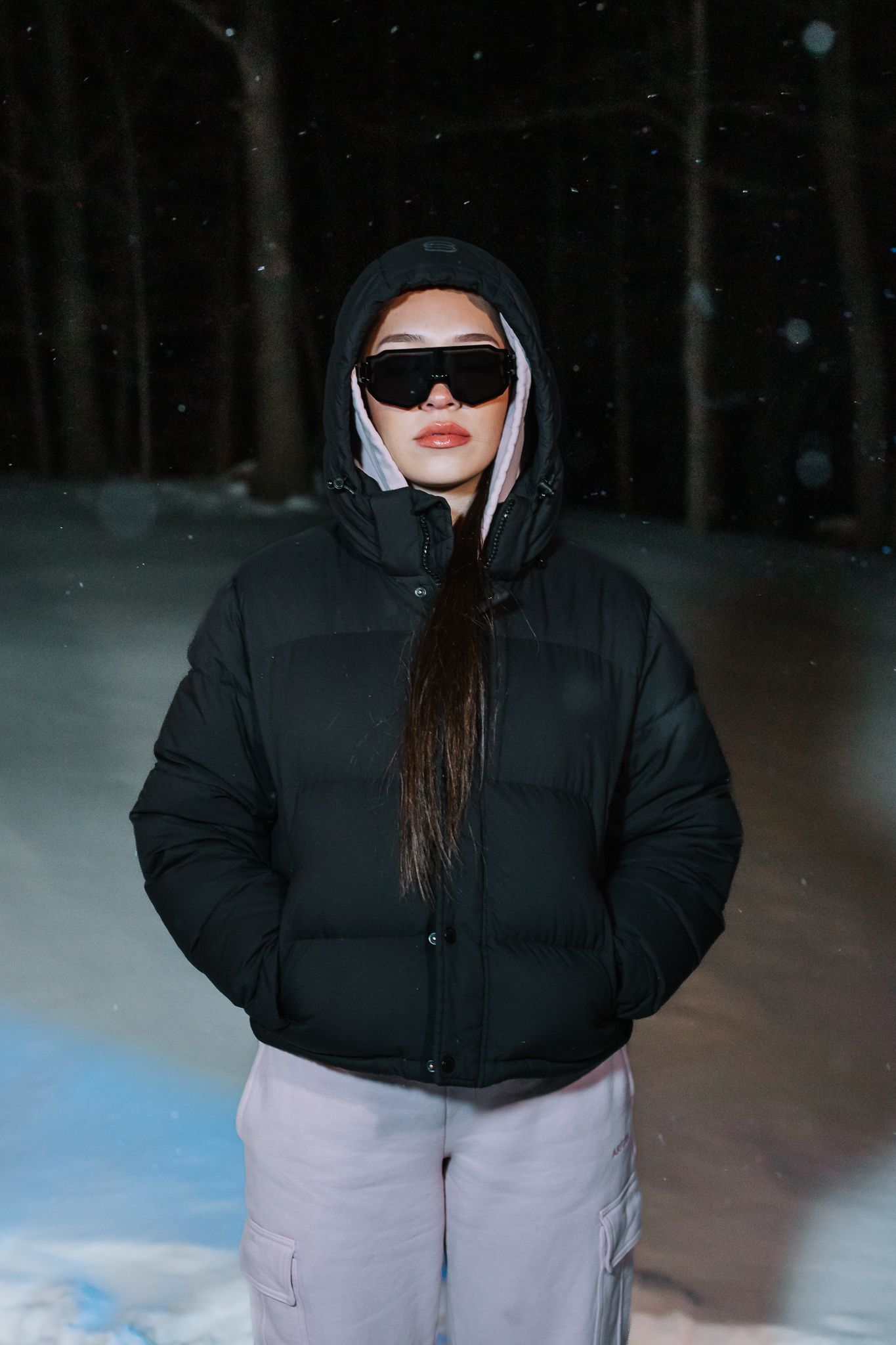 Woman in black puffer jacket with hood and black sunglasses standing in a snowy night forest.