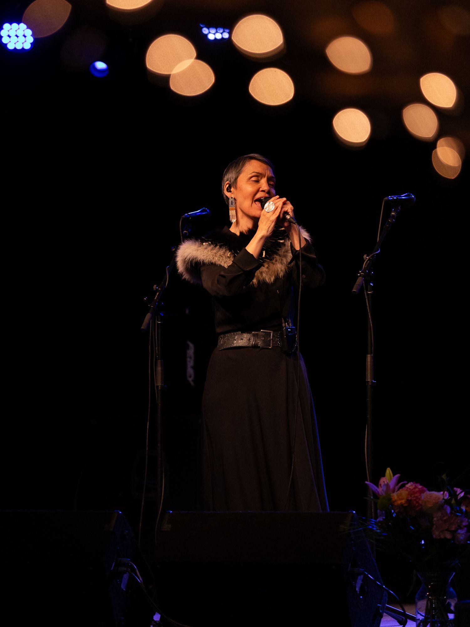 Woman in black dress with fur collar singing on stage with bokeh lights in background.