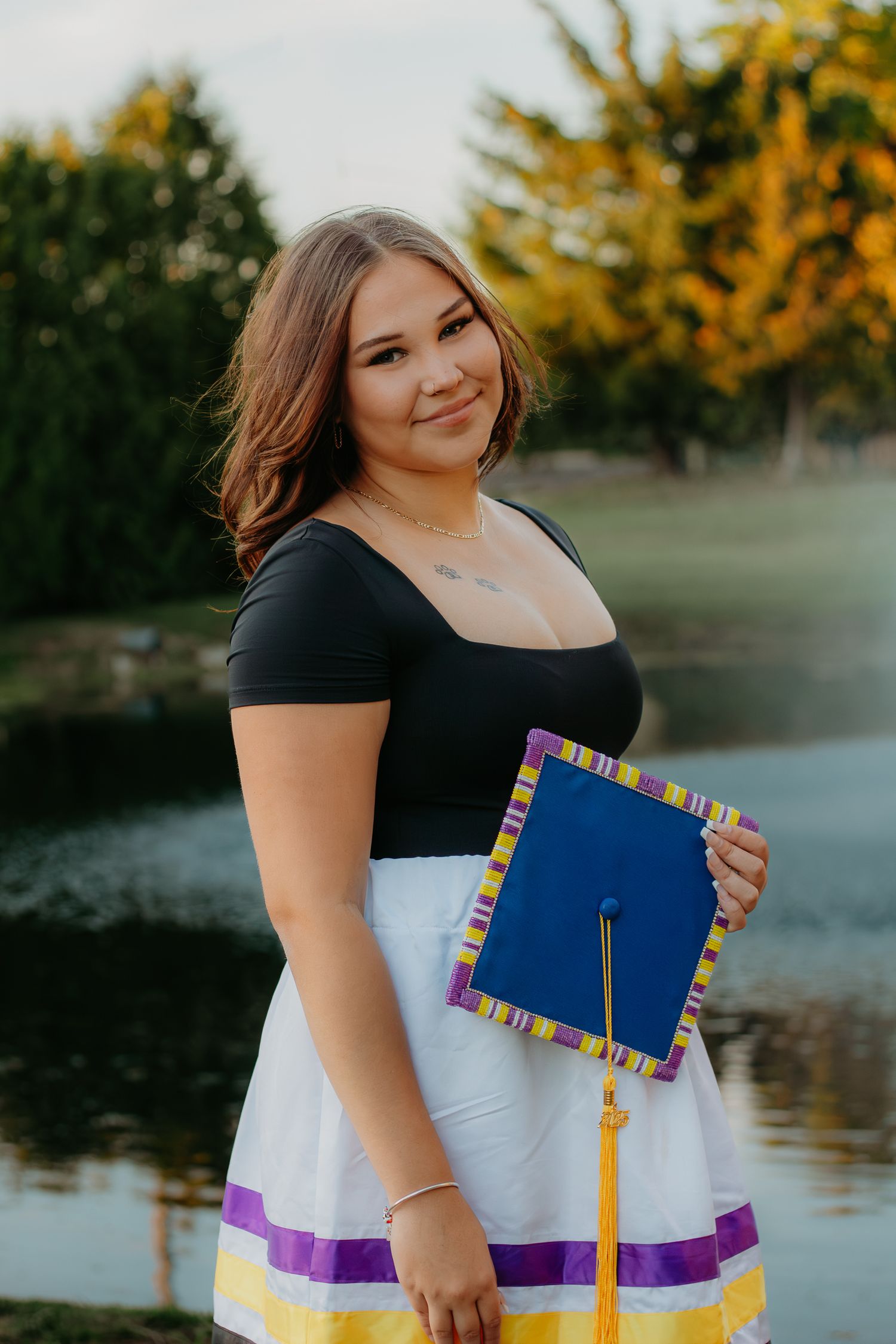 Graduate holding decorated cap poses by lake in black and white dress.