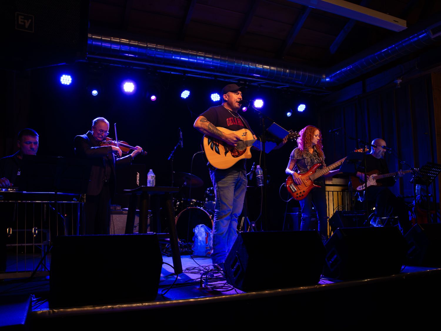 A band performs on stage with blue lighting, featuring guitar and violin players.