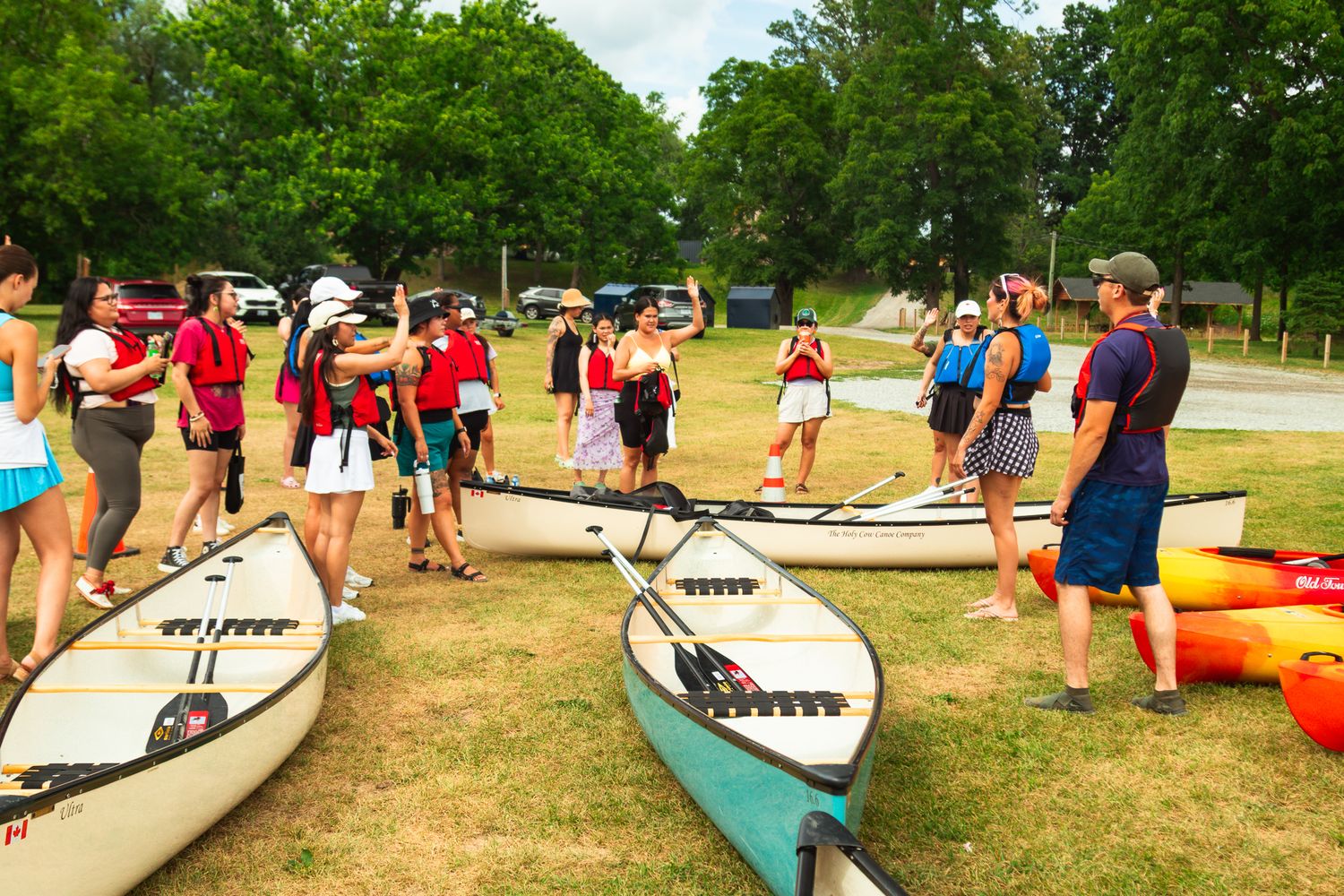 Group of people gathering around canoes and kayaks on grassy shore preparing for water activities.