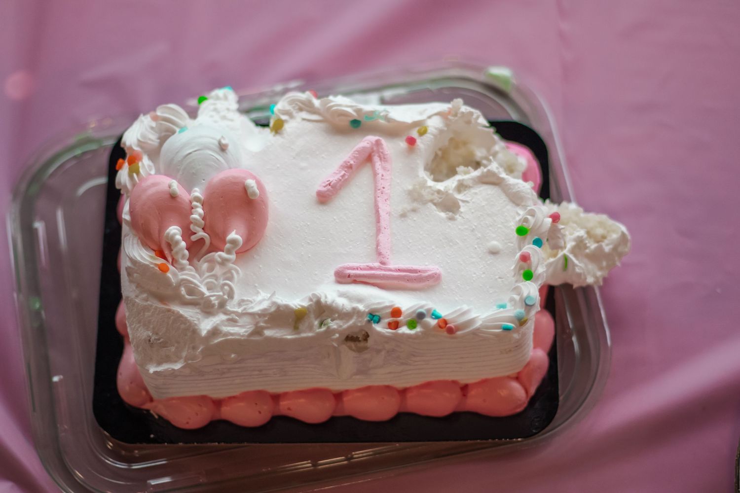 White and pink first birthday cake with number 1 candle, colorful sprinkles, and balloon decorations on pink tablecloth.