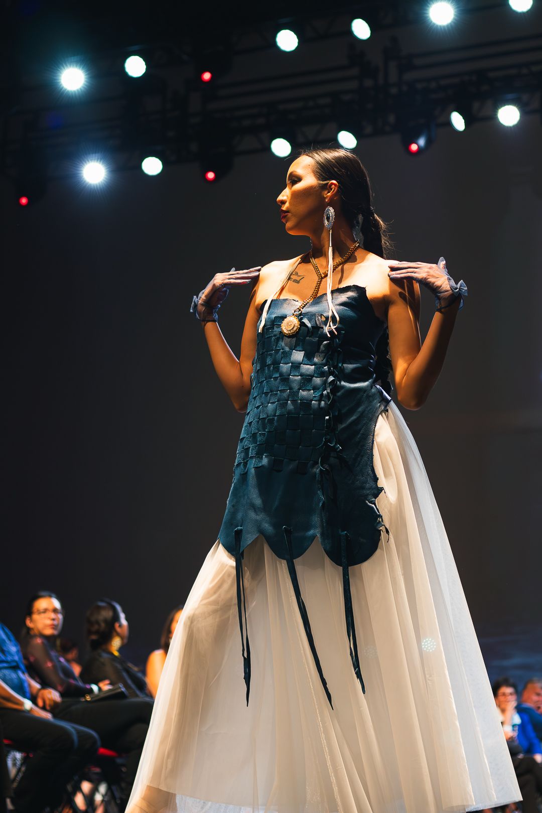 Model walks runway in blue woven bodice gown with white skirt at fashion show.