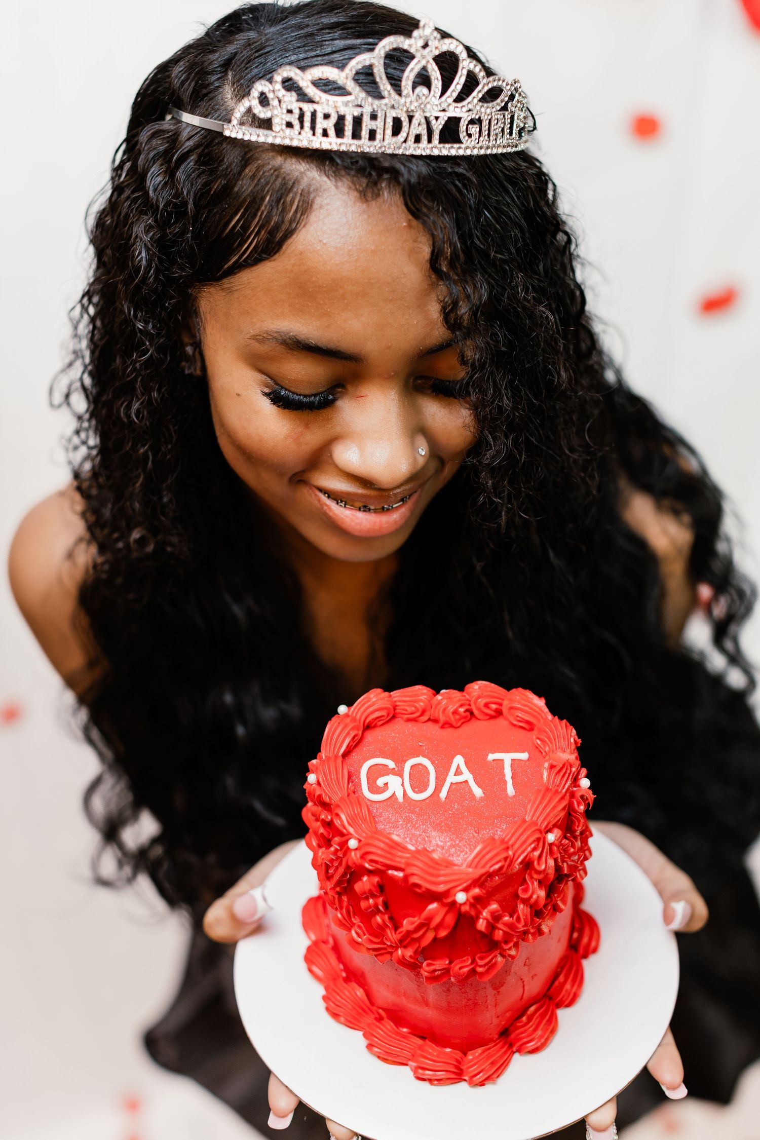 Young woman wearing a Birthday Girl tiara smiles at a red heart-shaped cake inscribed with GOAT.