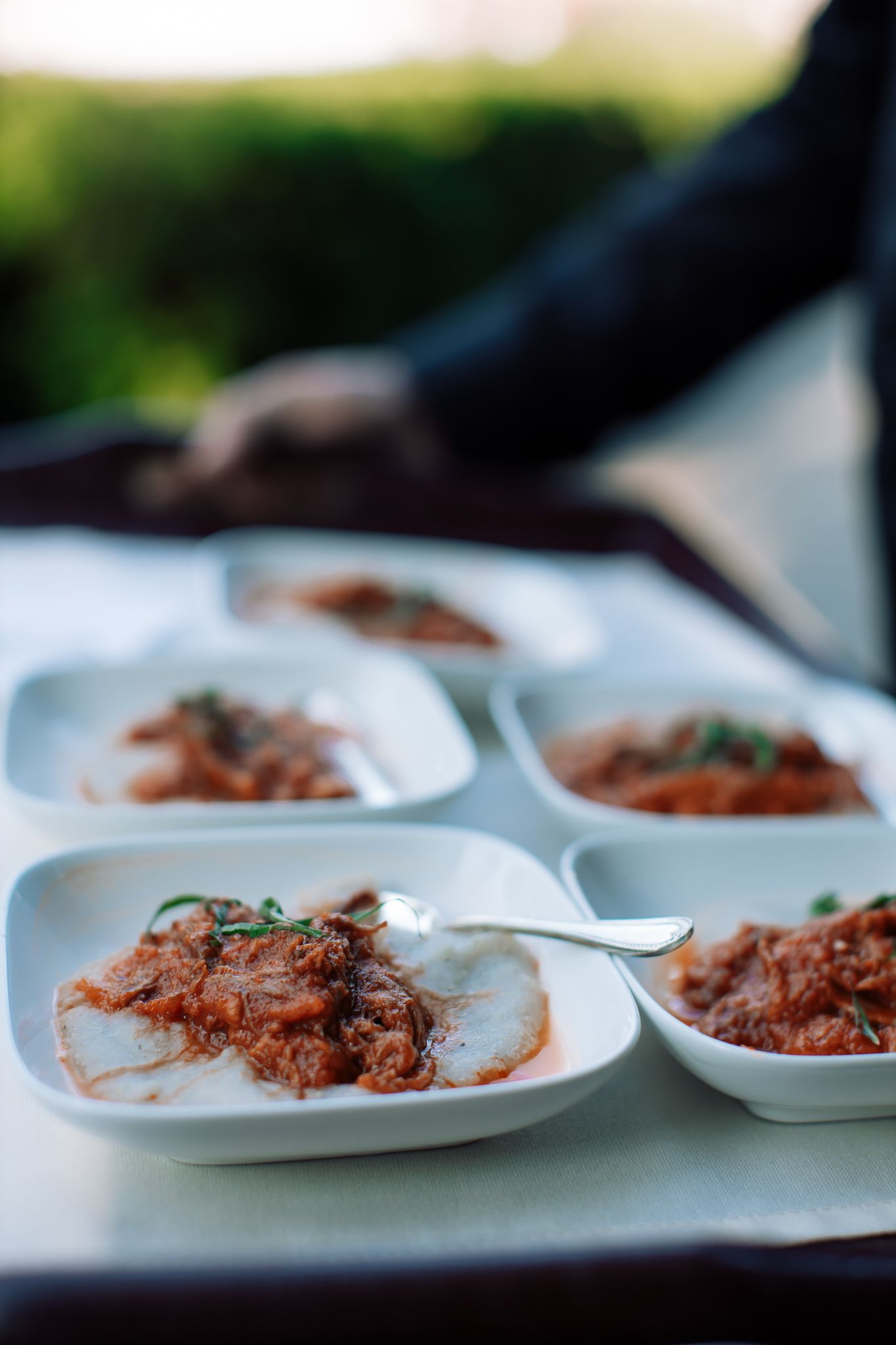 White square dishes of tomato-based stew garnished with herbs served on outdoor catering tray.