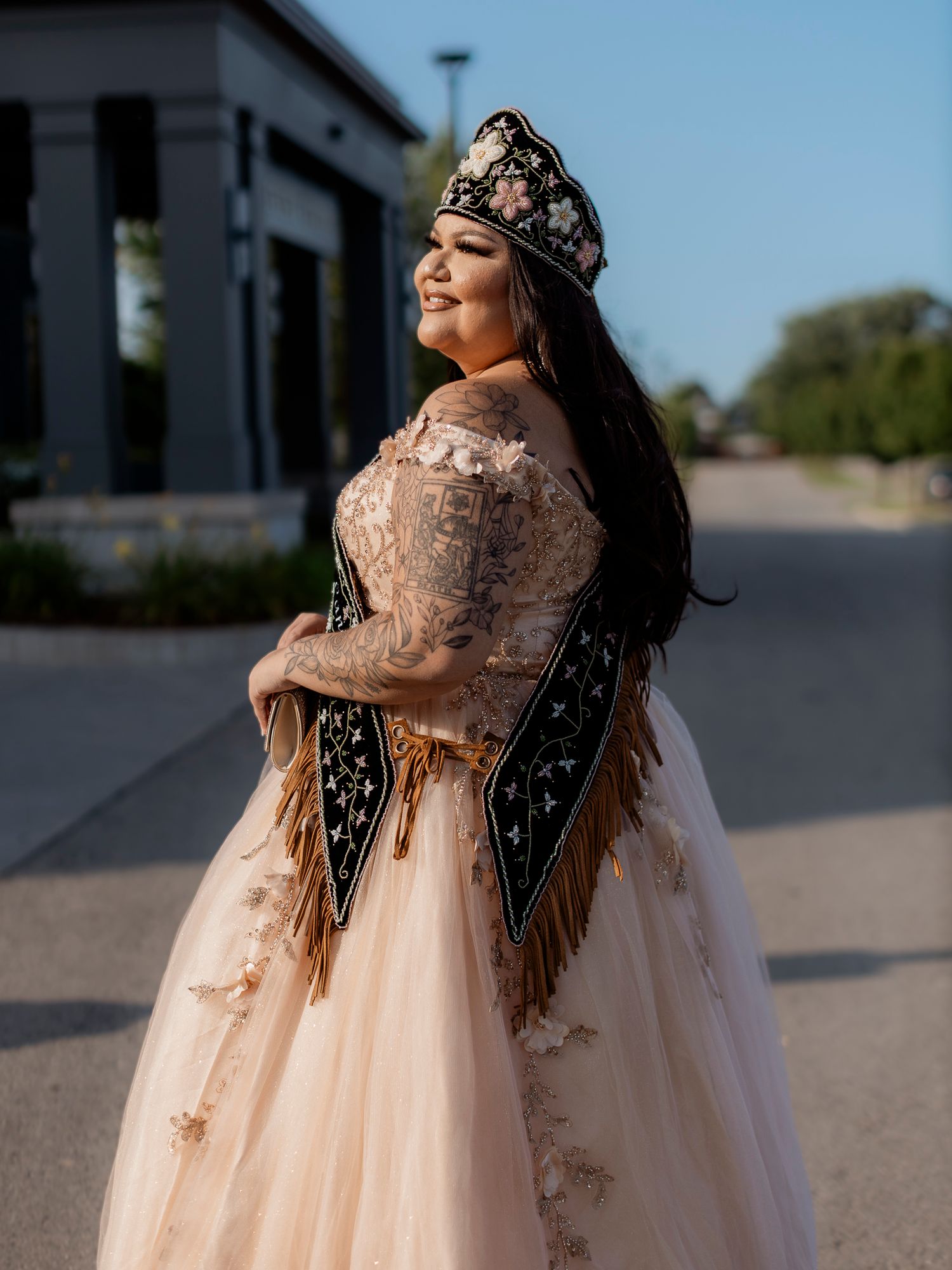 Young woman in elegant quinceañera gown with floral crown and embroidered sash posing outdoors.