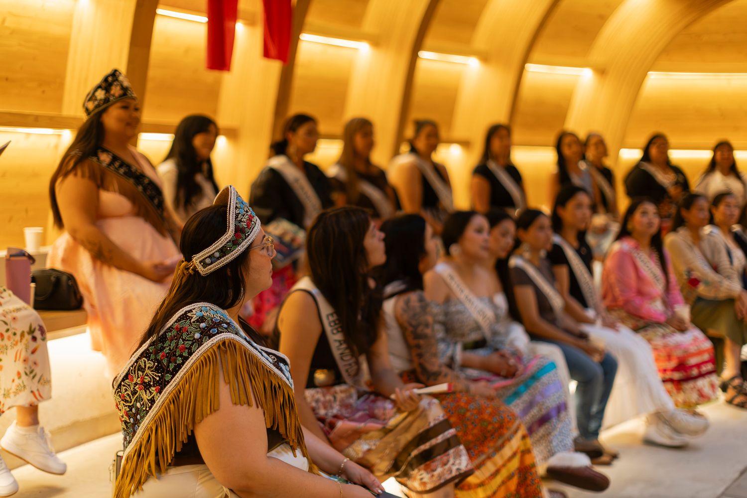 Native American women in traditional regalia seated during a cultural ceremony inside a warmly lit venue.