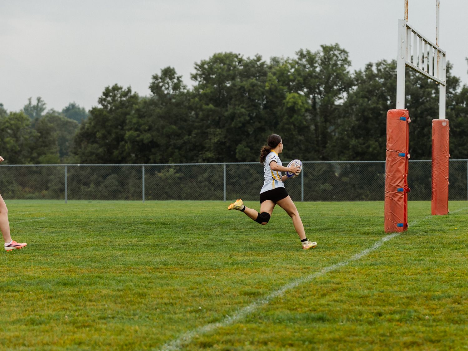 Female rugby player sprints toward the goal post carrying the ball during outdoor match.