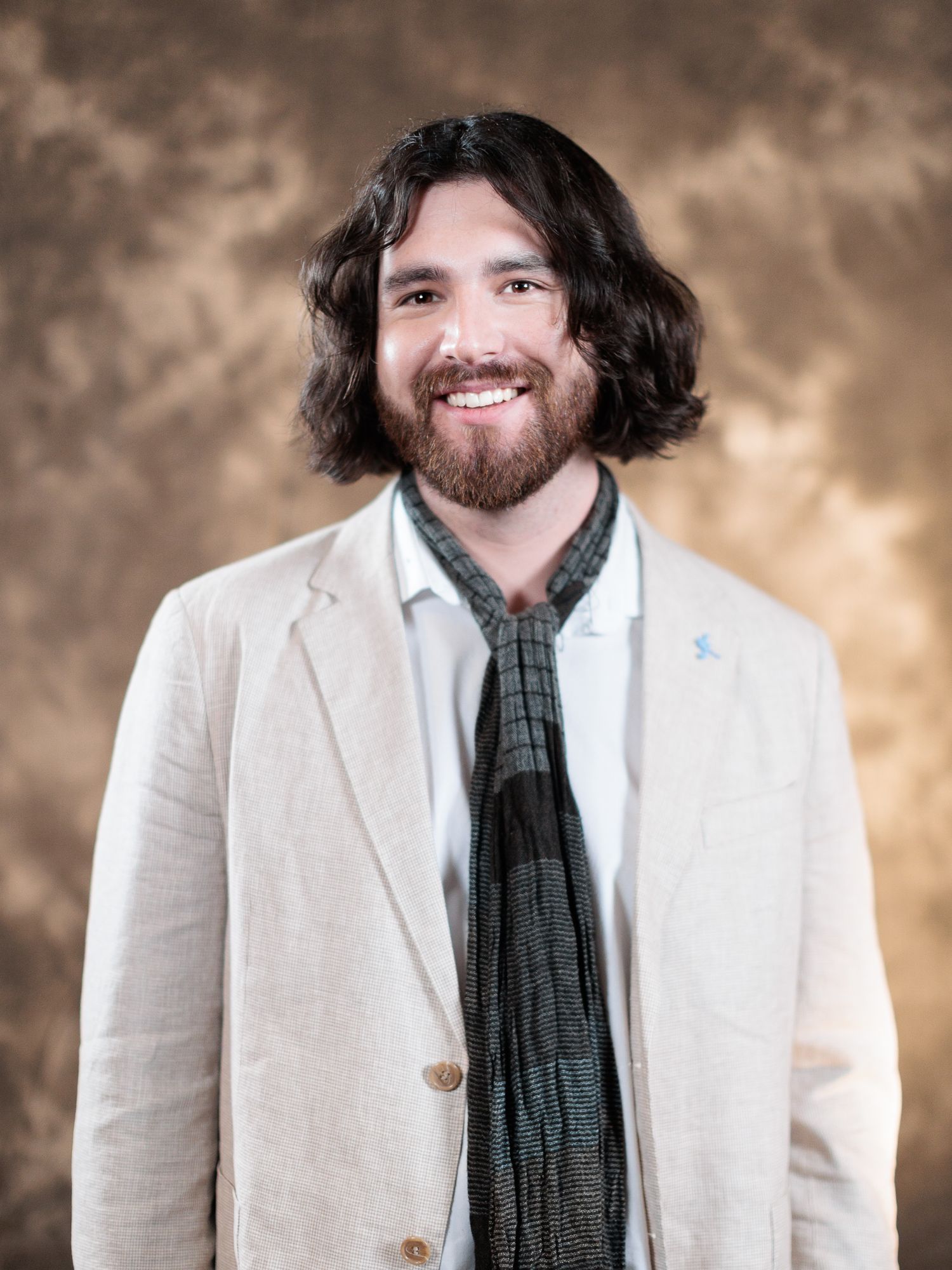 Smiling man with shoulder-length dark hair and beard wearing a white blazer and dark scarf against a brown backdrop.
