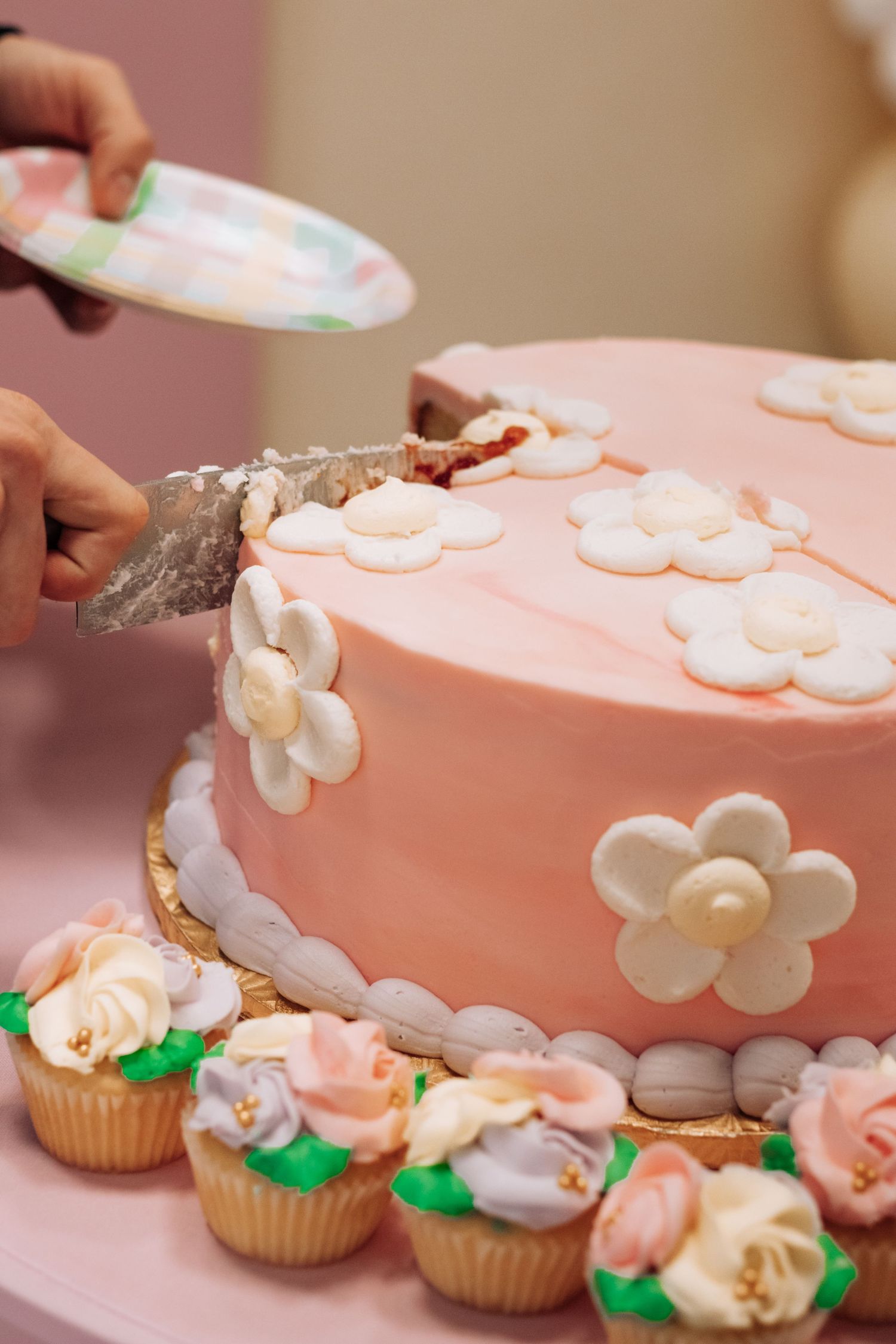 Hands cutting a pink fondant cake decorated with white flowers, surrounded by floral cupcakes at a celebration.