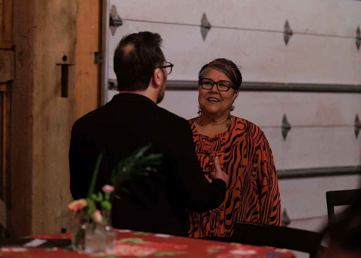 Two people engaged in conversation at an indoor event, woman in orange zebra-print top gestures while speaking to man in black jacket.