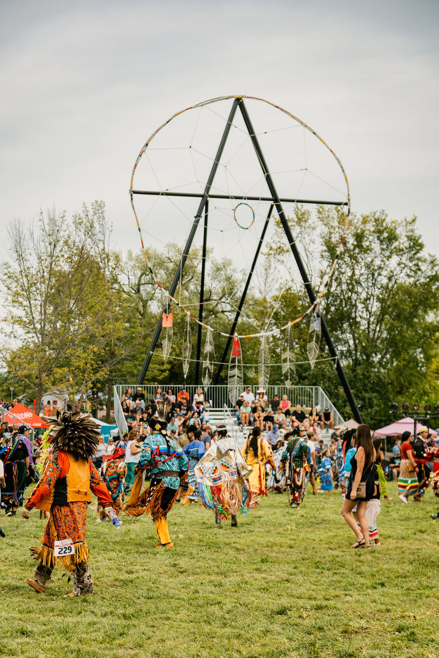 Native American dancers performing in a circle under tall wooden poles at an outdoor powwow.