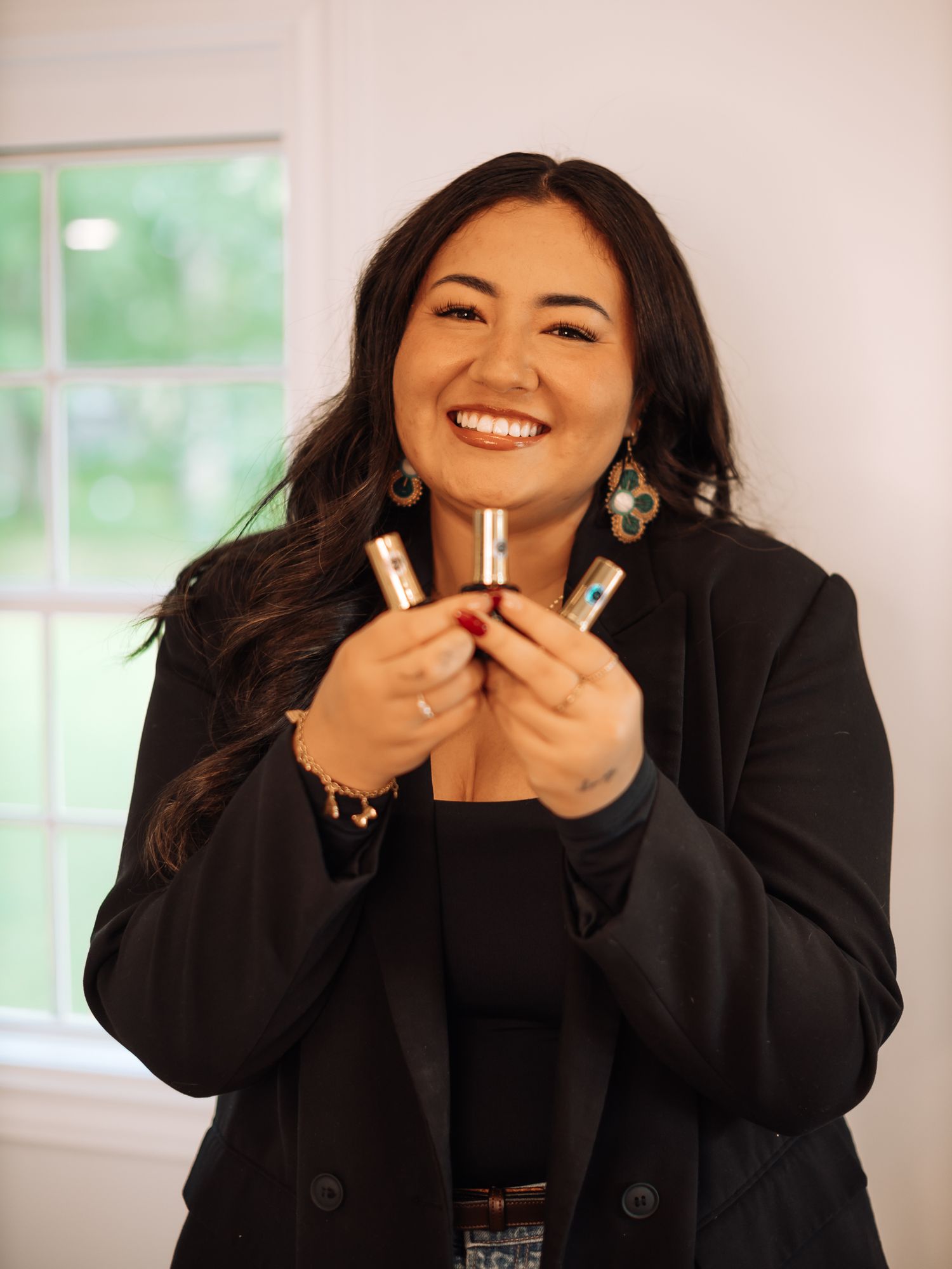 A person in a black blazer holds up small items while smiling in front of a window.