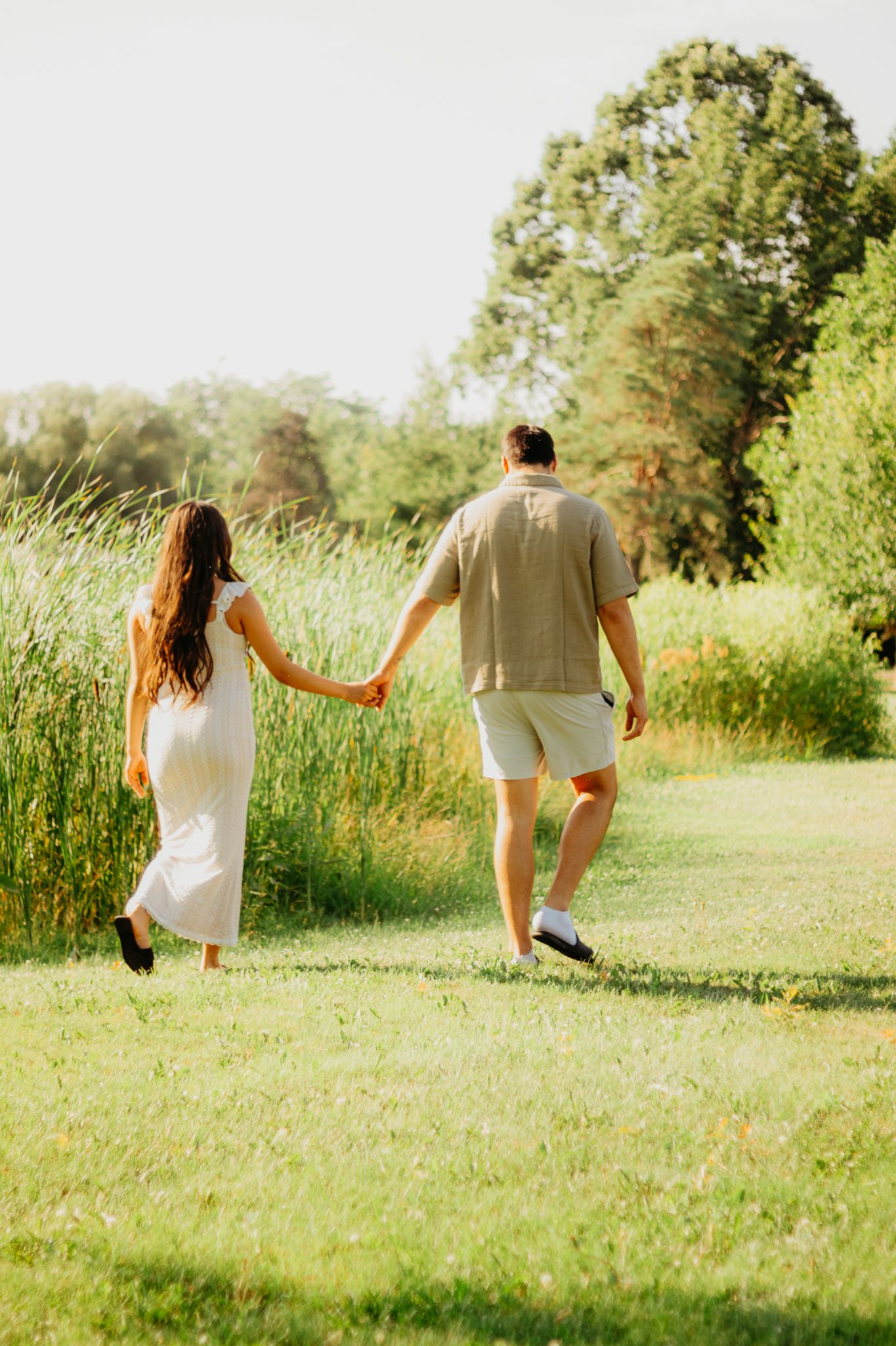 A couple runs and plays together on a grassy path bordered by tall reeds in golden sunlight.