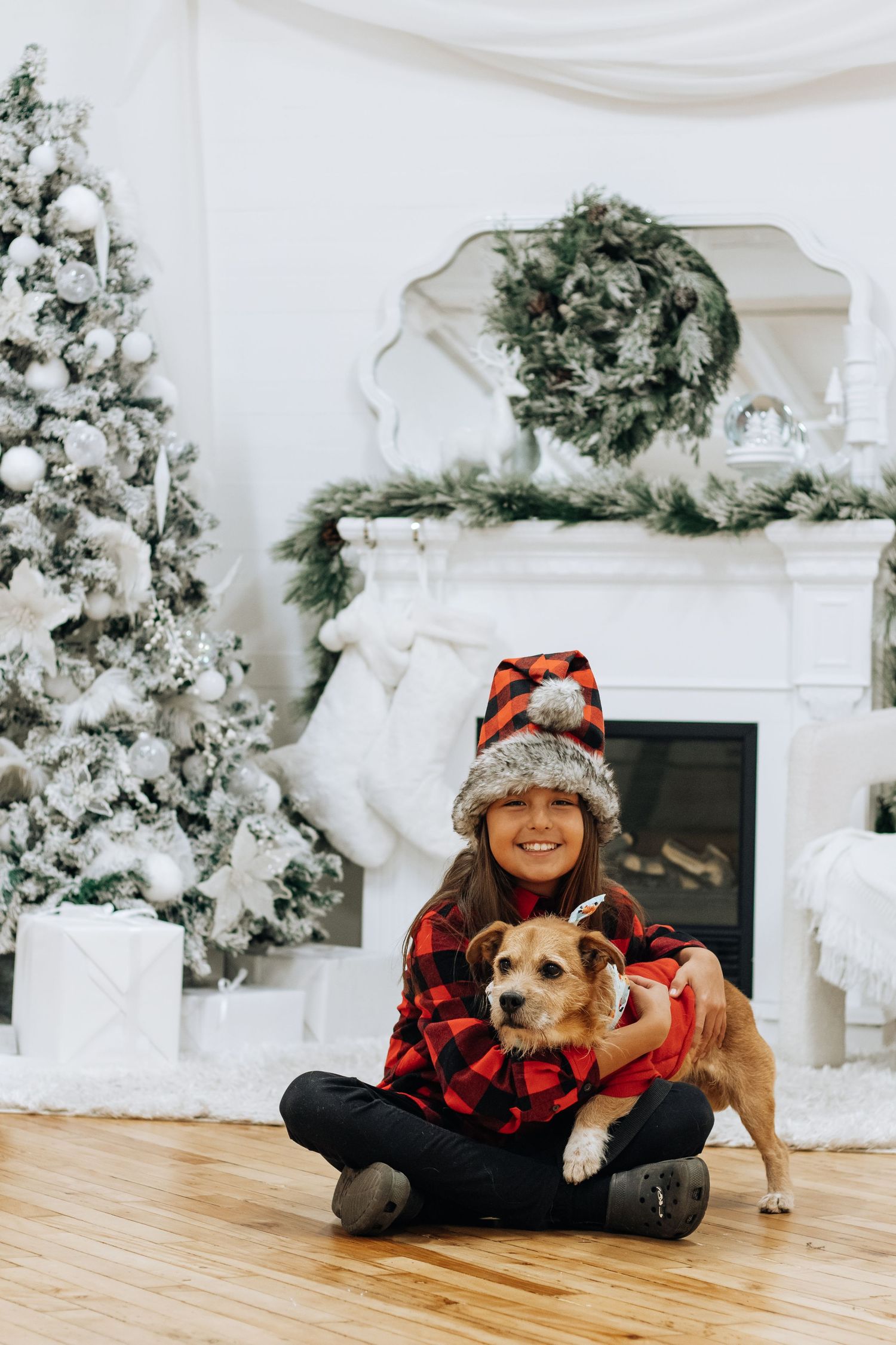 Person in buffalo plaid cuddling with dog on wooden floor near Christmas decorations.