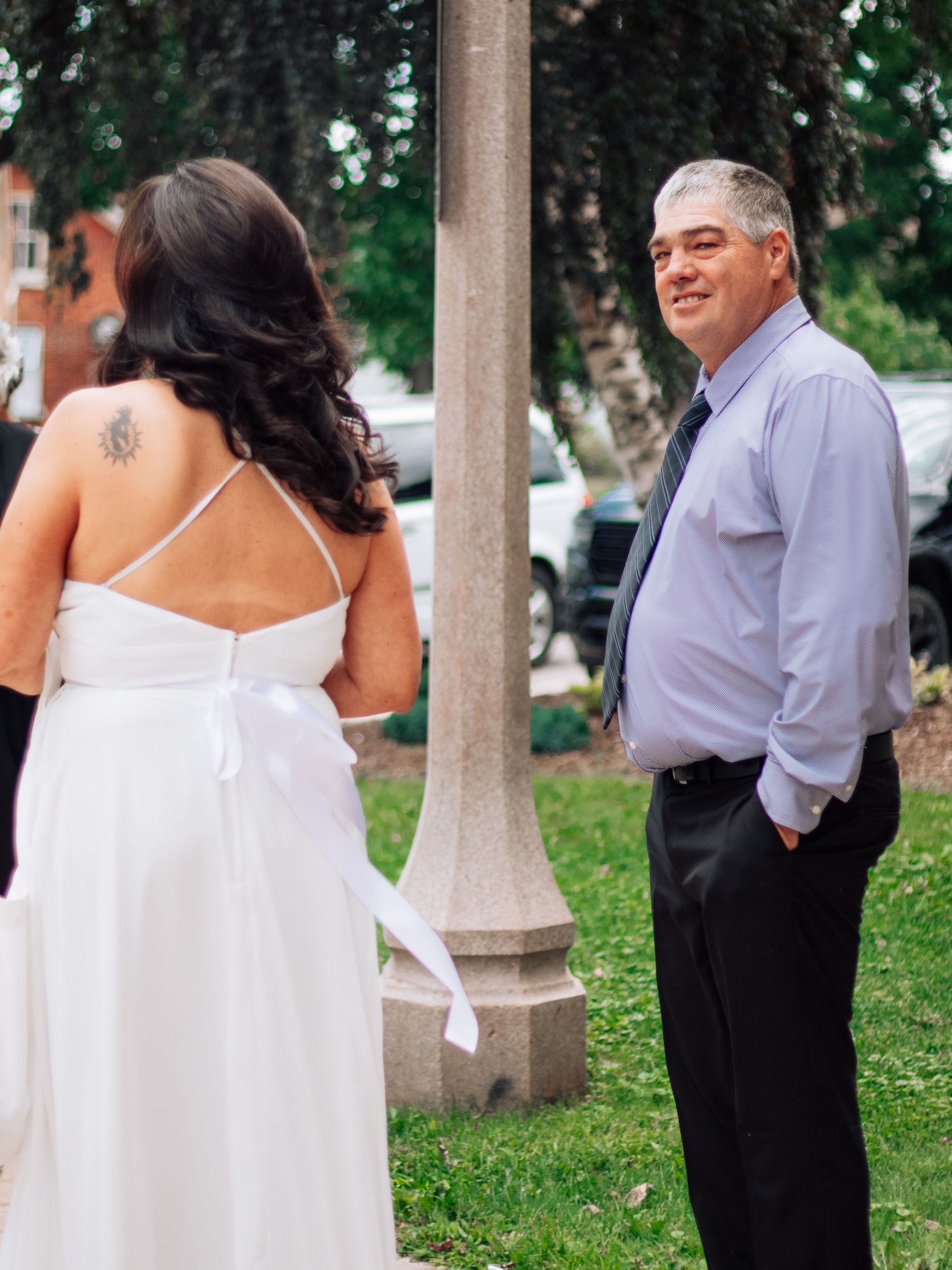 Groom in blue dress shirt and tie smiles at bride in backless white wedding gown near a lamppost outdoors.