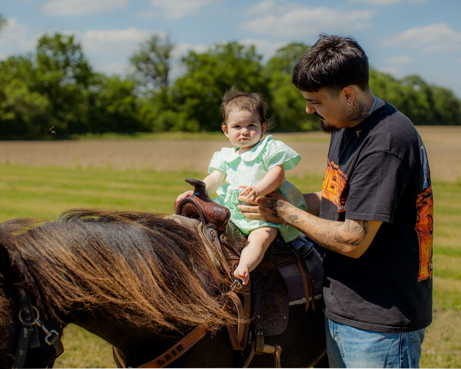 A man helps a baby girl sit on a dark horse in a sunny green field at a rodeo-themed birthday party.