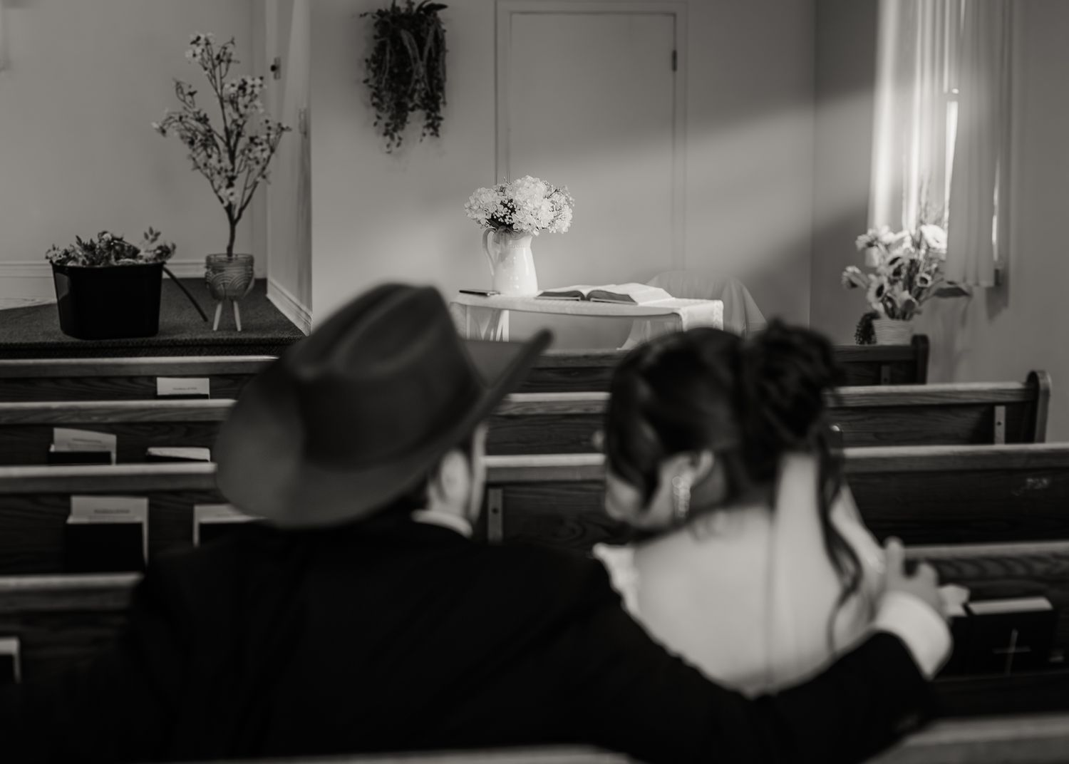 Black and white photo of a couple sitting together in church pews, facing a floral altar.