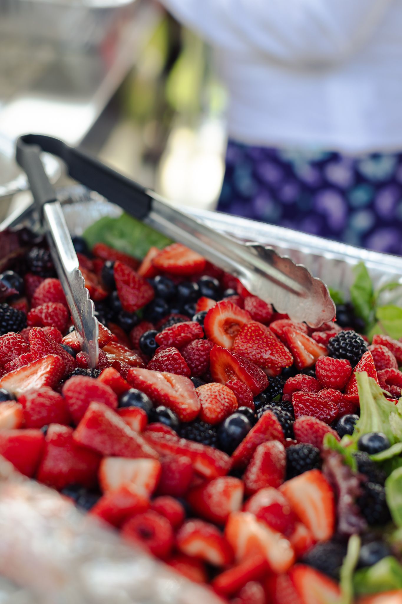 Fresh mixed berry platter with strawberries, raspberries, and blueberries served with tongs at a catered outdoor event.