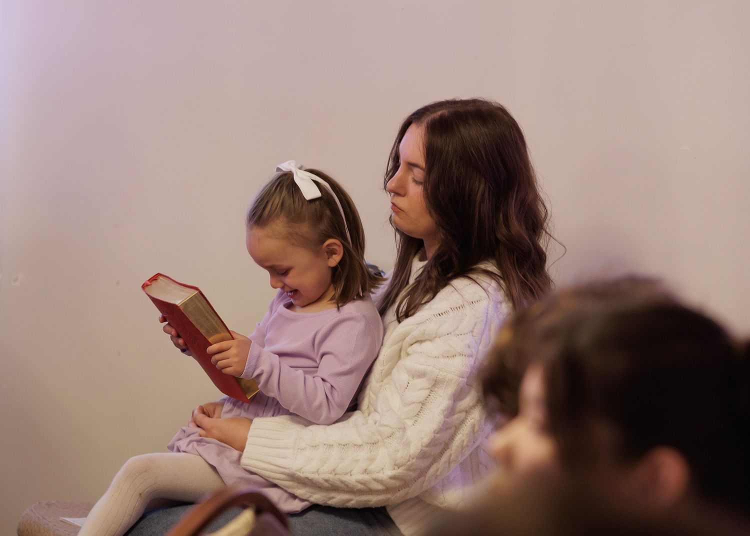 A mother and young daughter sit together reading a red book, the girl in a purple dress with a white bow.