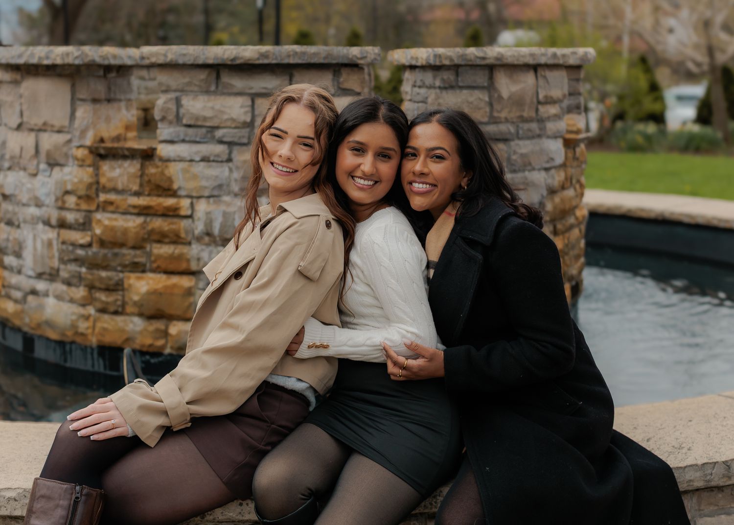 Three young women sitting together outdoors, smiling and embracing near a stone fountain.