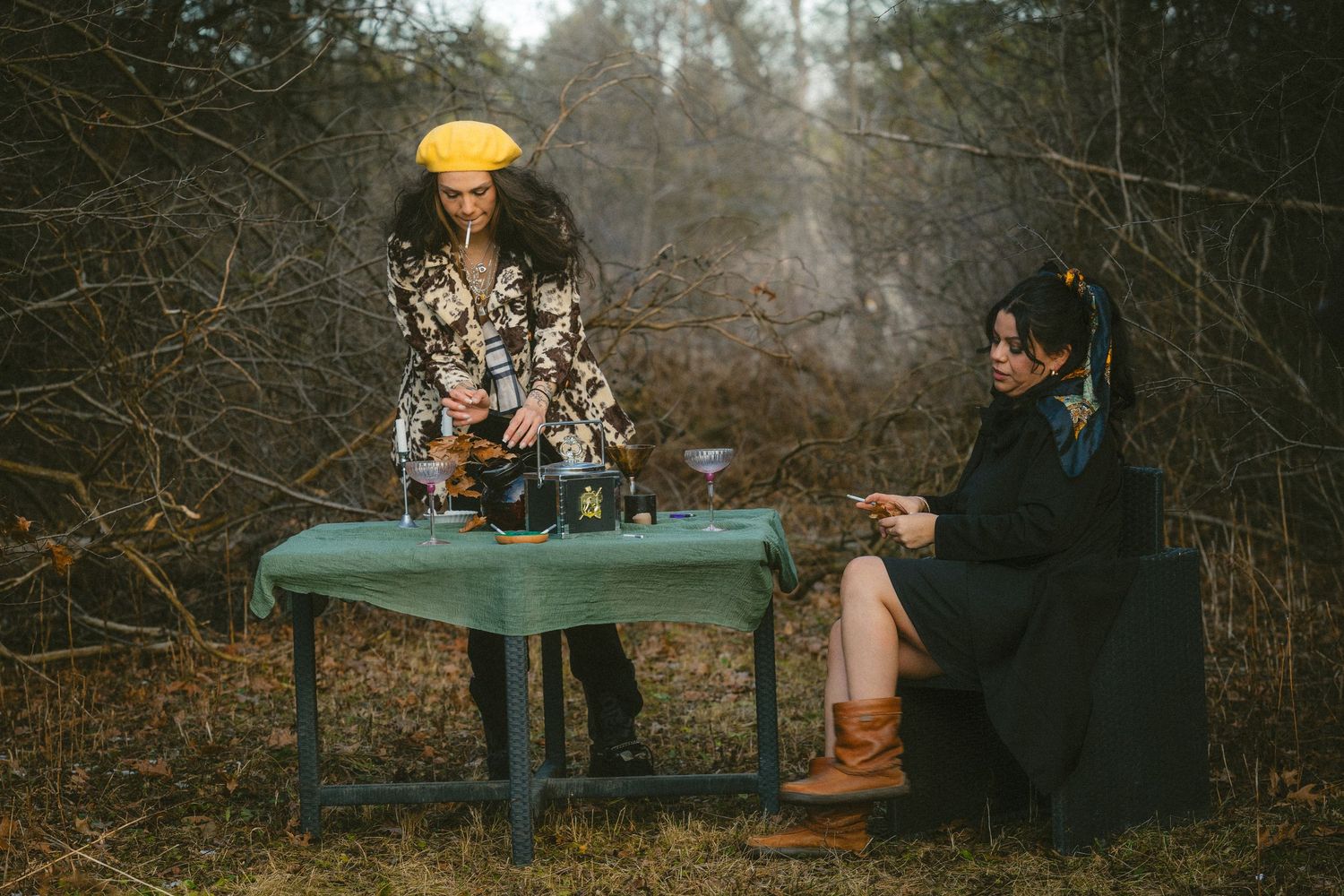 Two women in bohemian outfits share tea at a rustic outdoor table surrounded by bare winter trees.