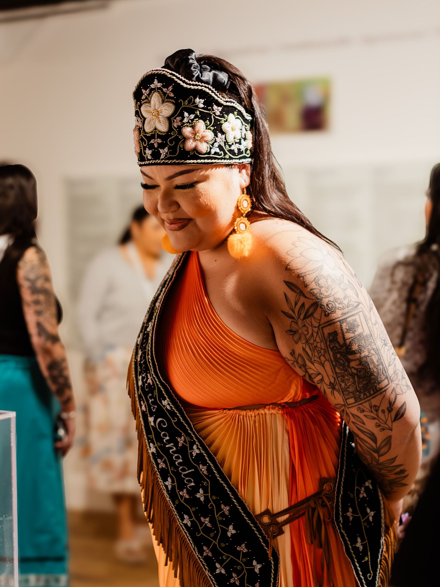 Woman in orange dress and black embroidered headdress with fringe and tattoos smiles.
