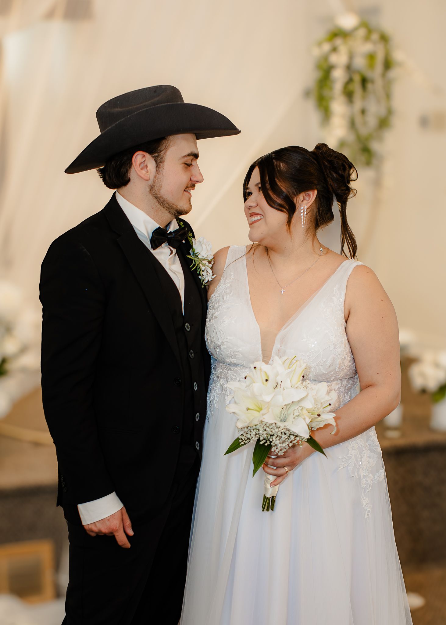 Groom in black cowboy hat and suit gazes at bride holding white lily bouquet in lace wedding gown.