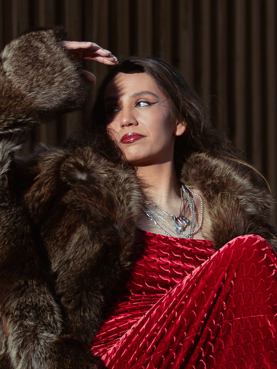 Woman in red velvet dress and fur coat with silver jewelry, shielding eyes from sunlight.