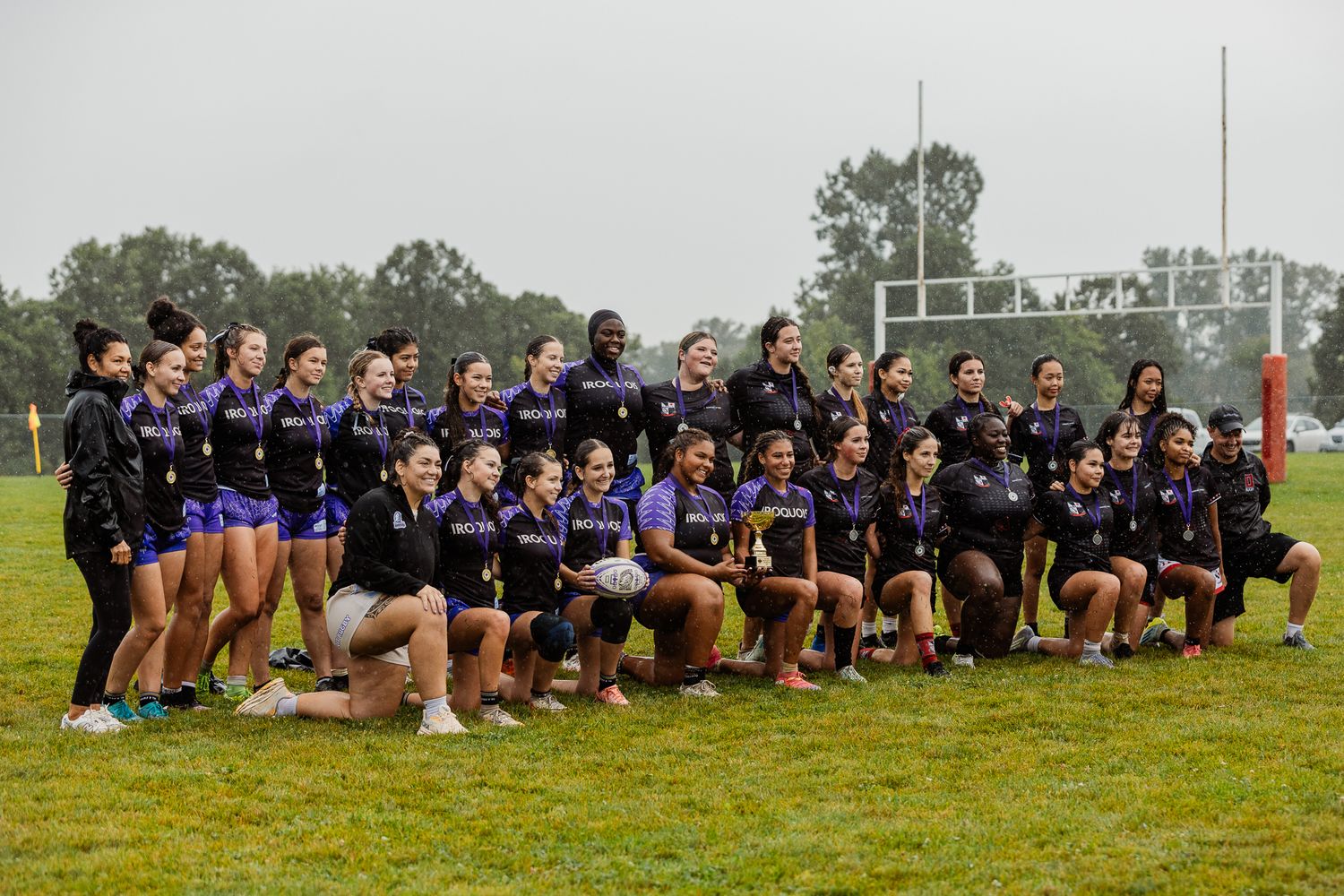 Women’s rugby team poses together on a rainy field in purple jerseys for a group photo.