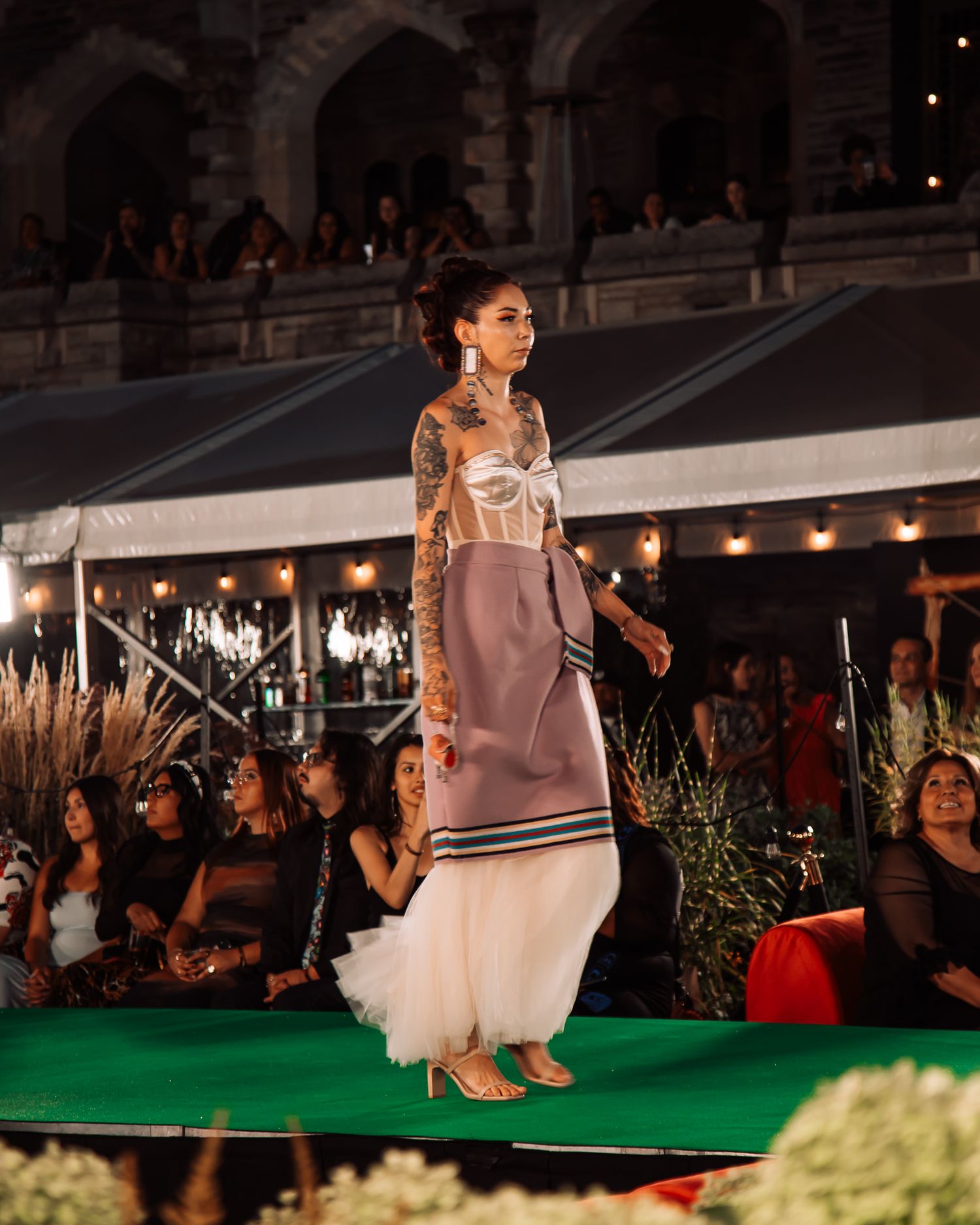 A tattooed model walks the runway in a elegant mauve skirt and corset top with white tulle hem at an outdoor fashion show.