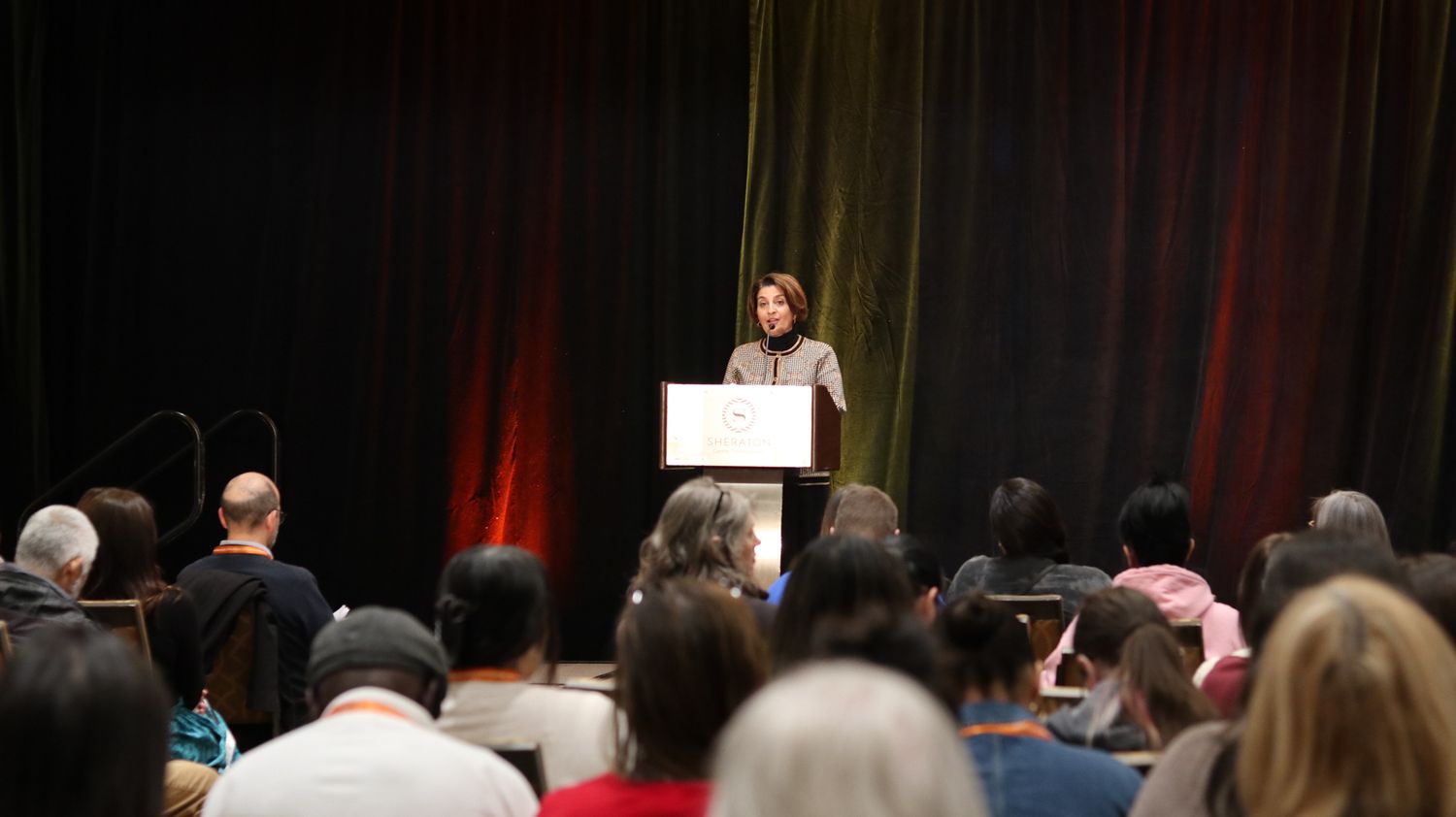 Woman speaking at podium on stage before a large seated audience at a conference.
