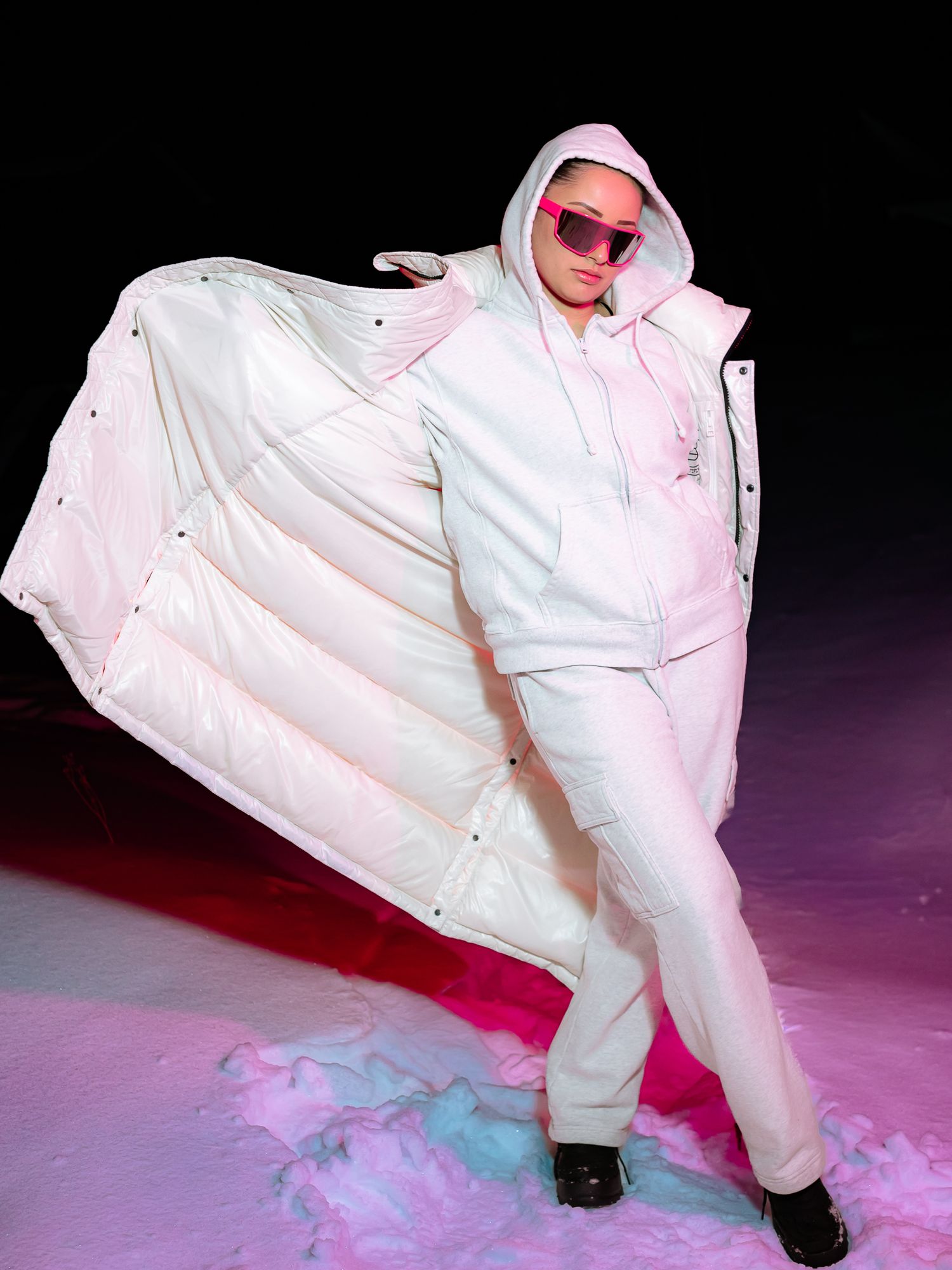 Model in all-white puffer coat and matching outfit with pink sunglasses posing dynamically on snowy surface.