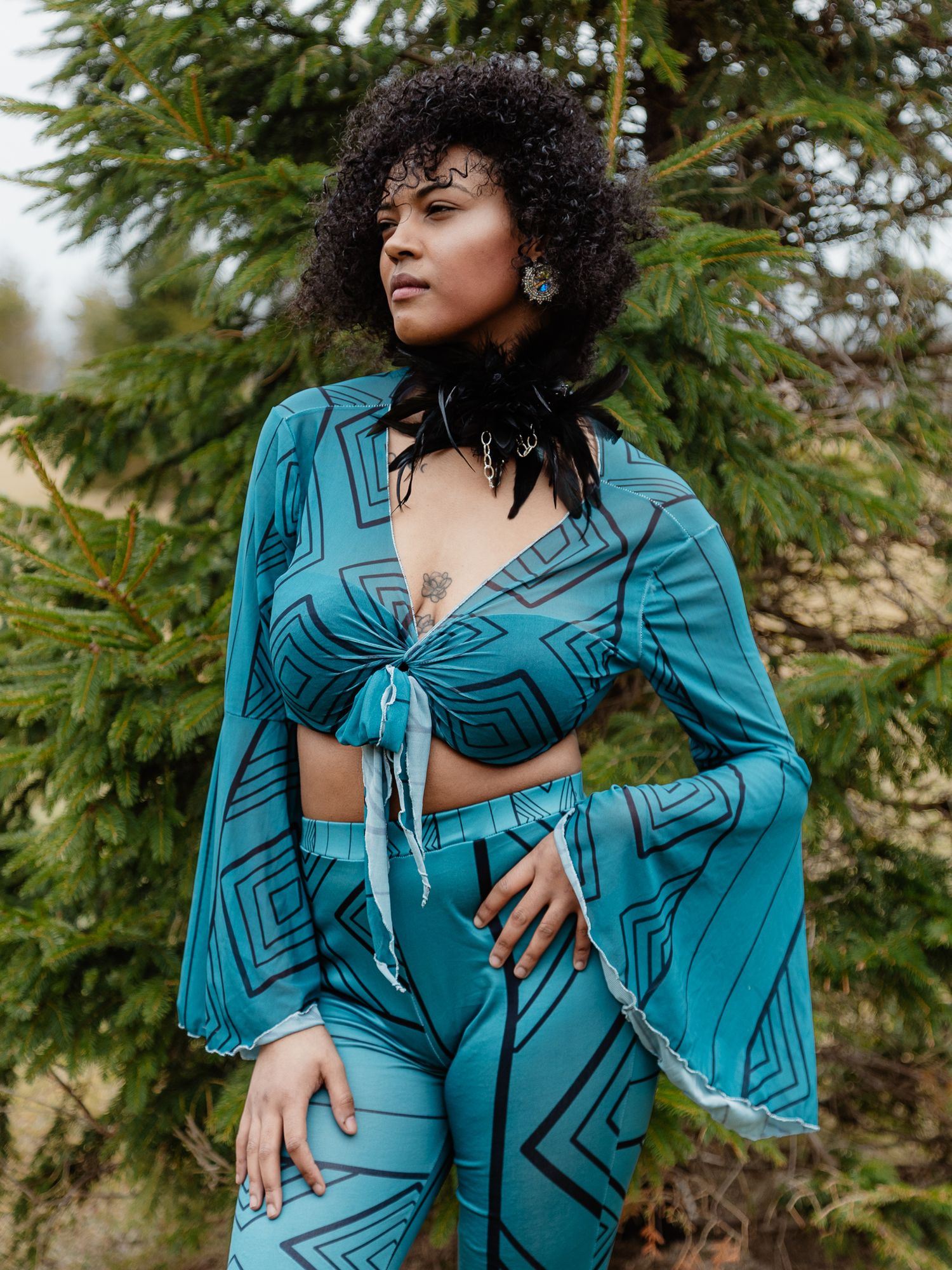 Woman in teal geometric print matching crop top and flare pants set, posing outdoors near evergreen trees.