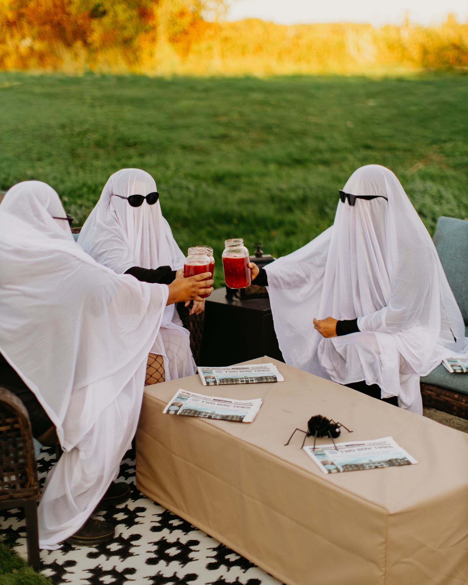 Two figures in ghost costumes and sunglasses toast drinks at an outdoor table with newspapers.