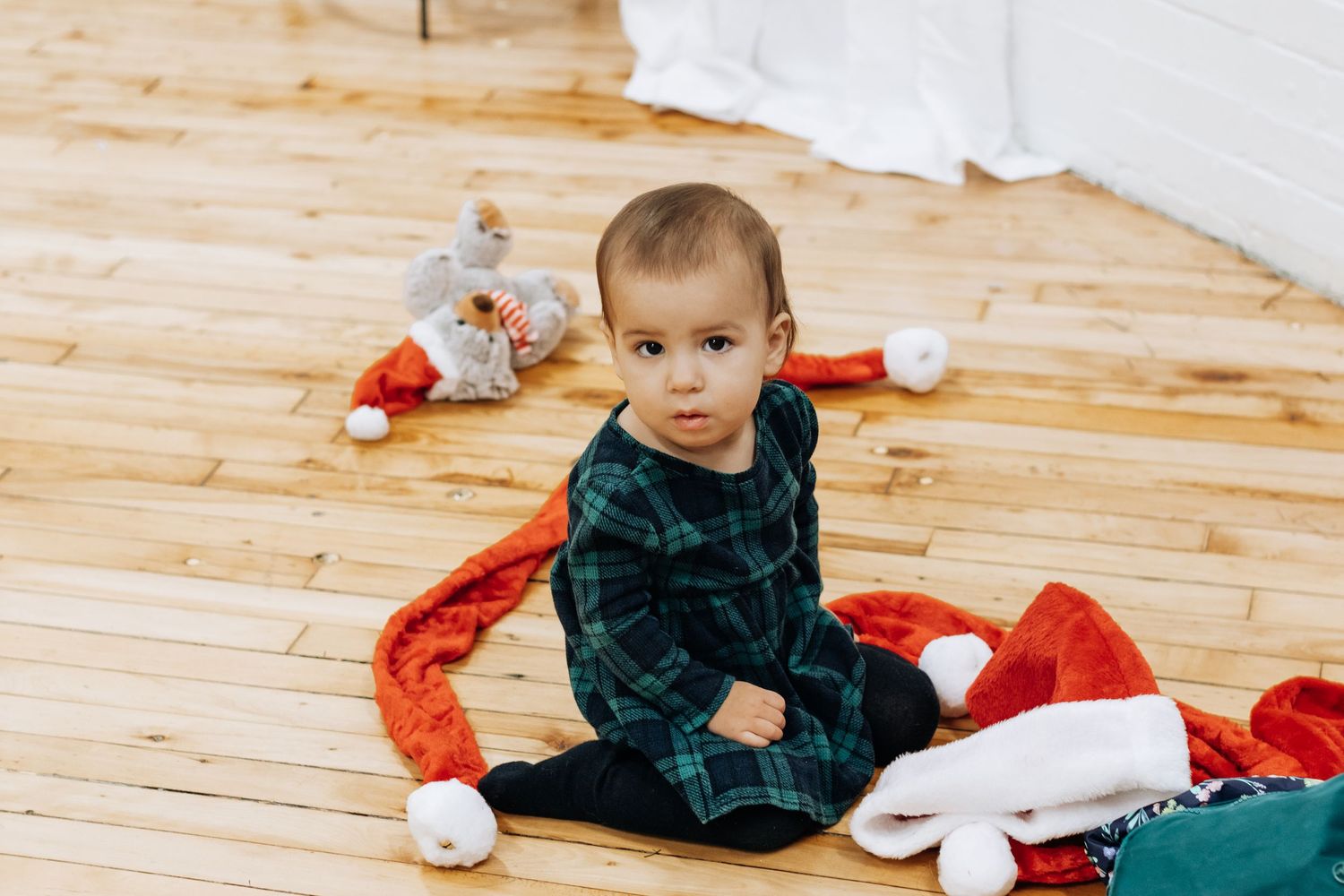Child in plaid shirt playing with red holiday decorations on wooden floor.