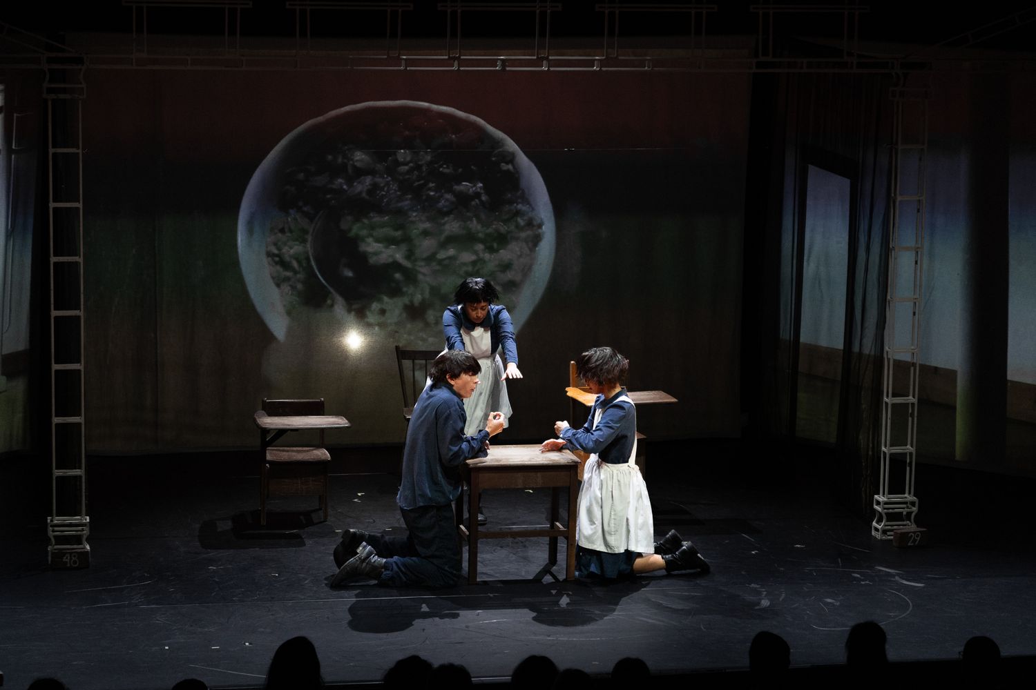 A dramatic stage performance scene with actors at a table under moody lighting and a large moon backdrop.