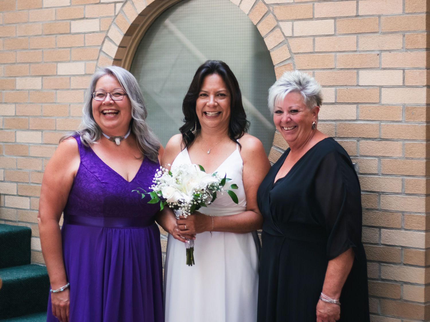 Bride in white gown holding bouquet poses with two women in purple and black dresses outside church.
