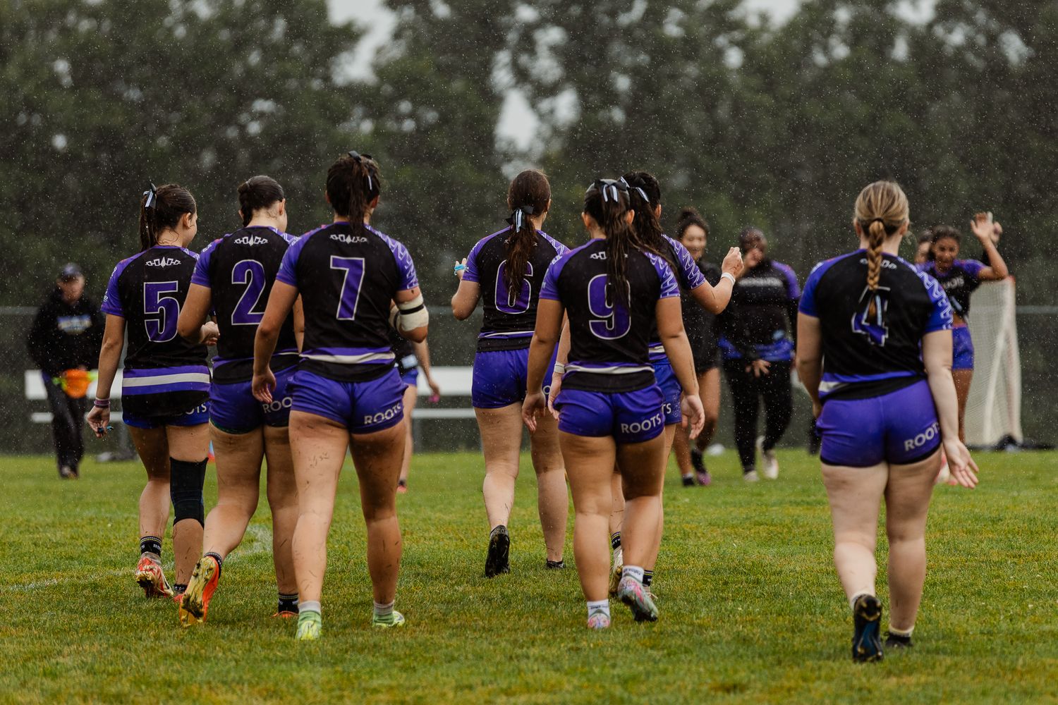 Female rugby players in purple uniforms walk across a rain-soaked field showing jersey numbers.