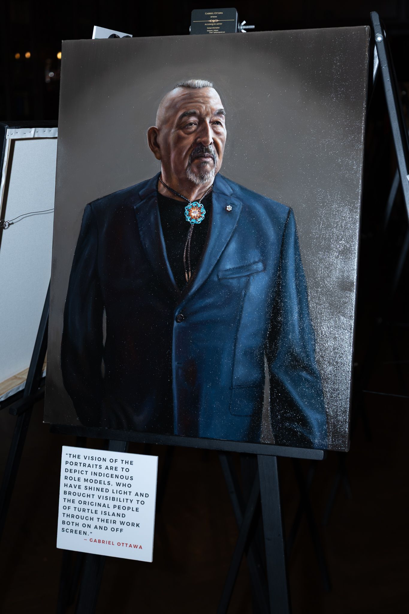 Portrait painting of an Indigenous elder in a blue suit with turquoise bolo tie displayed on an easel at an art exhibition.