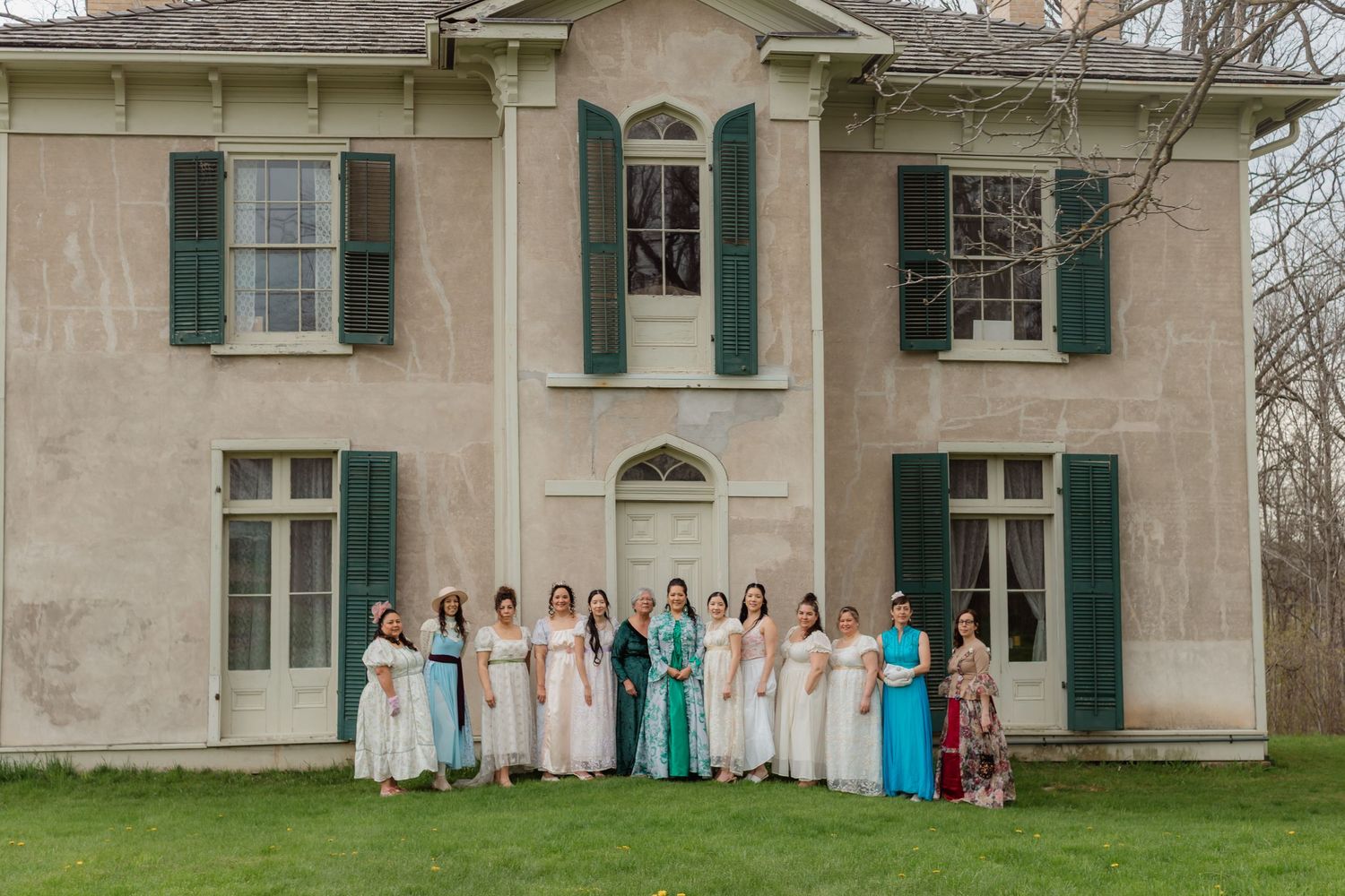 Group of women in formal dresses posing outside historic two-story stone house with green shutters.