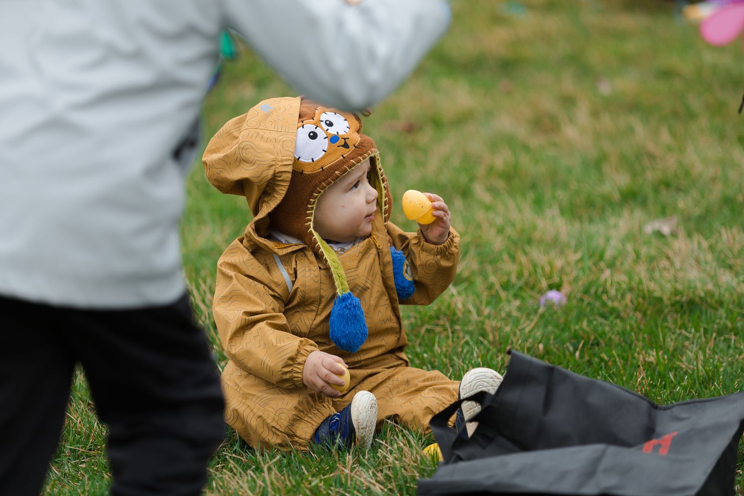 Toddler in brown bear costume sitting on grass eating at an outdoor Easter egg hunt event.