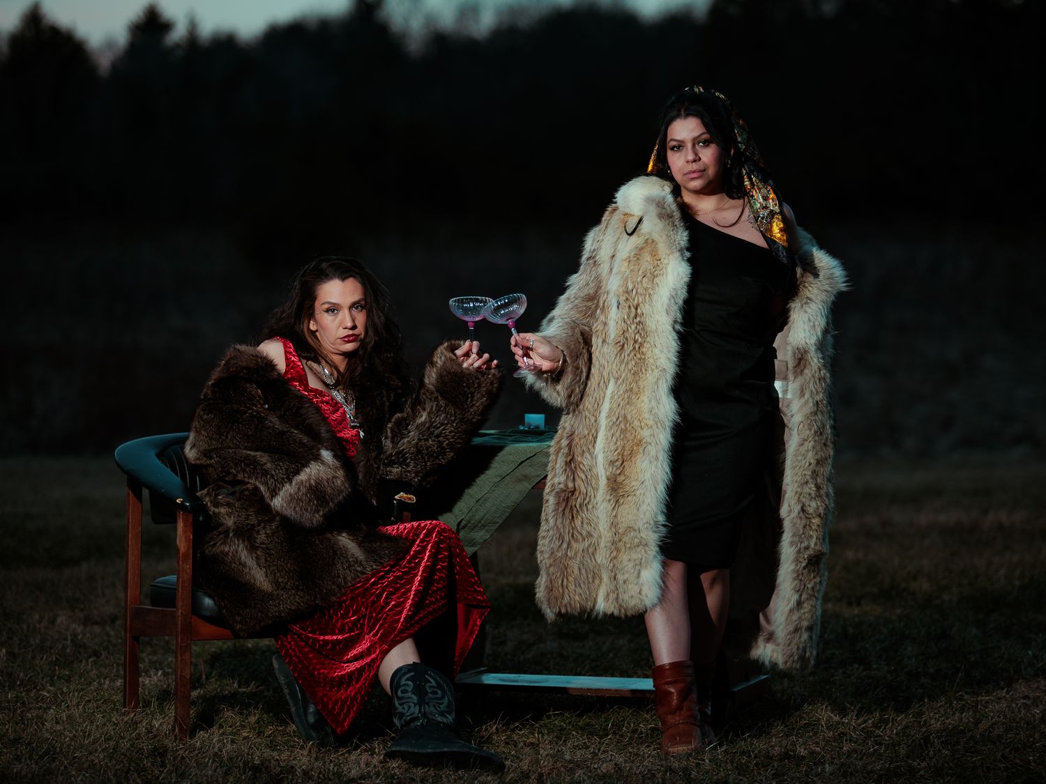 Two women in fur coats holding cocktail glasses outdoors at twilight.