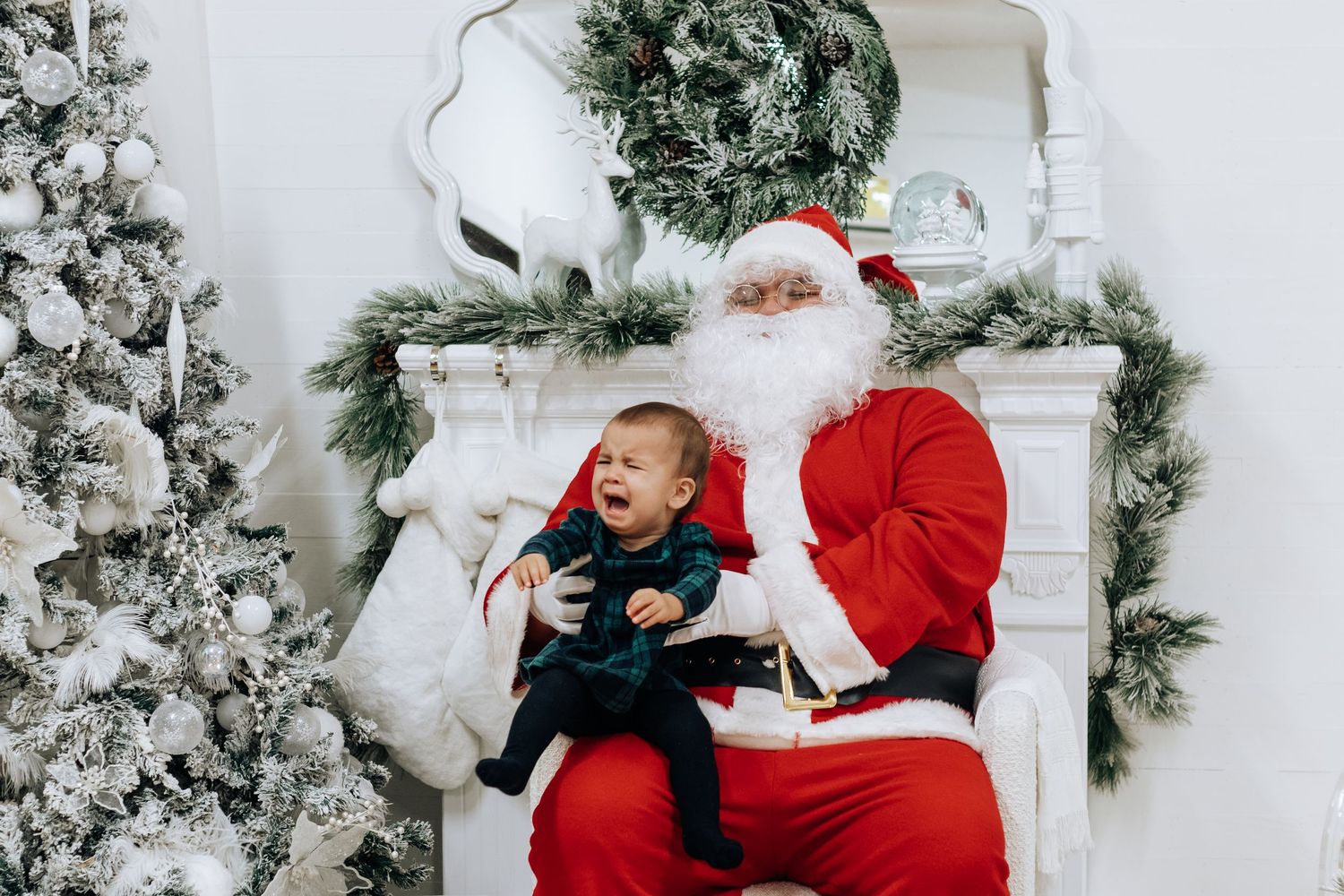 Holiday portrait session featuring Santa Claus and young child in front of snowy white Christmas decor.